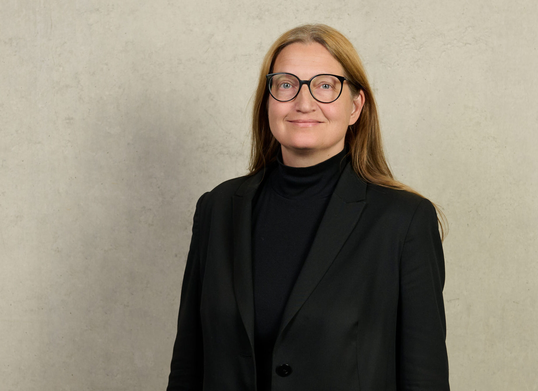 Eine Person mit langen hellbraunen Haaren und Brille trägt einen schwarzen Blazer und einen schwarzen Rollkragenpullover und steht vor einer schlichten hellen Wand für ein BR-Interview mit Prof. Meißner über offene Placebos. at Hochschule Coburg