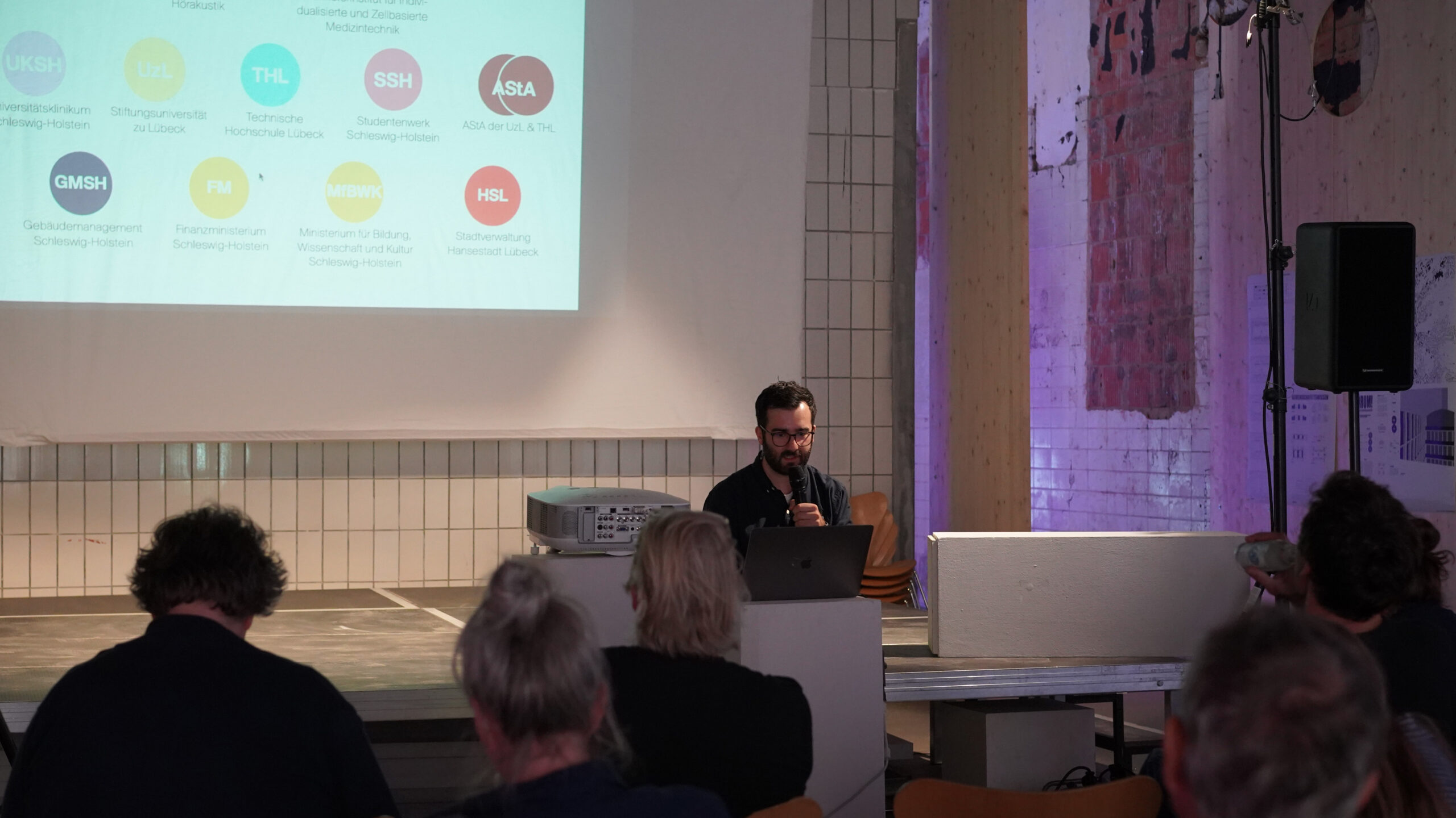 Ein Mann spricht in ein Mikrofon, während er vor einem Publikum an einem Laptop sitzt und über transformative Stadtentwicklung spricht. Auf einer Projektionsfläche hinter ihm sind bunte kreisförmige Logos und Texte zu sehen. Der Raum ist modern und im industriellen Stil eingerichtet. at Hochschule Coburg