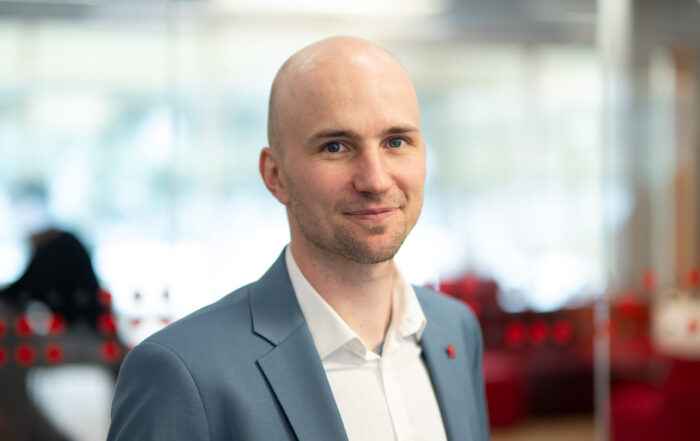 Ein Mann mit rasiertem Kopf, hellblauem Blazer und weißem Hemd steht in einem modernen, hell erleuchteten Büro, das von der Innovation der Smart Factories inspiriert ist, mit verschwommenem roten und weißen Dekor im Hintergrund. at Hochschule Coburg