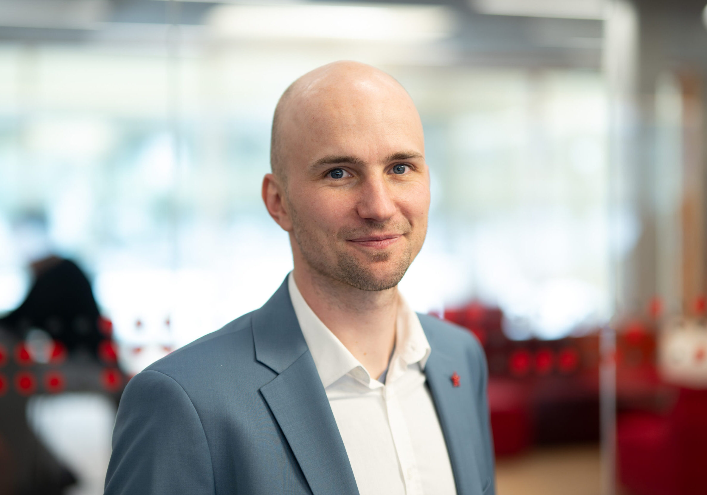 Ein Mann mit rasiertem Kopf, hellblauem Blazer und weißem Hemd steht in einem modernen, hell erleuchteten Büro, das von der Innovation der Smart Factories inspiriert ist, mit verschwommenem roten und weißen Dekor im Hintergrund. at Hochschule Coburg