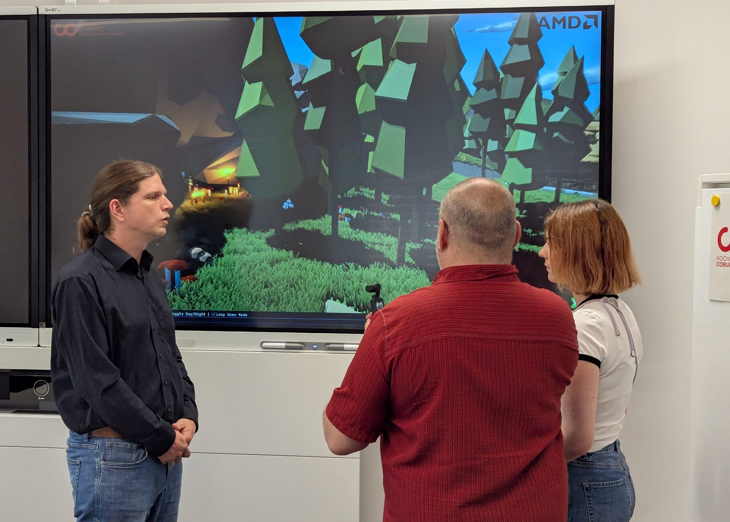 Drei Personen stehen vor einem großen Bildschirm, der eine stilisierte Videospielszene im Wald zeigt. Eine Person wendet sich der Gruppe zu, während die beiden anderen den Bildschirm beobachten und miteinander diskutieren. at Hochschule Coburg