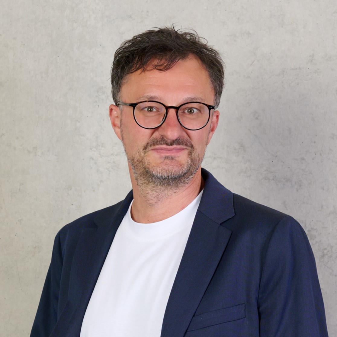 Ein Mann mit kurzen dunklen Haaren, Brille und gestutztem Bart trägt einen marineblauen Blazer über einem weißen T-Shirt und steht vor einer schlichten hellgrauen Betonwand. at Hochschule Coburg