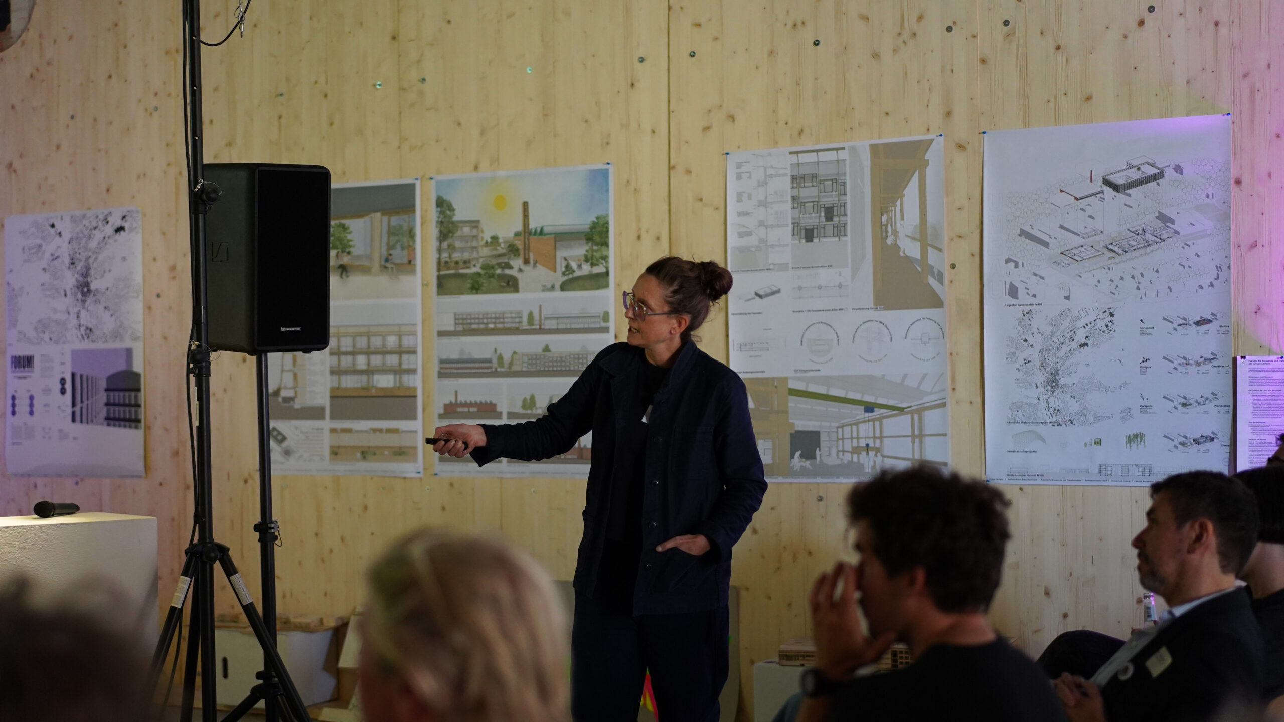 Eine Person steht und gestikuliert, während sie einem sitzenden Publikum in einem Raum mit Holzwänden architektonische Pläne auf Plakaten präsentiert und über transformative Stadtentwicklungskonzepte diskutiert. at Hochschule Coburg