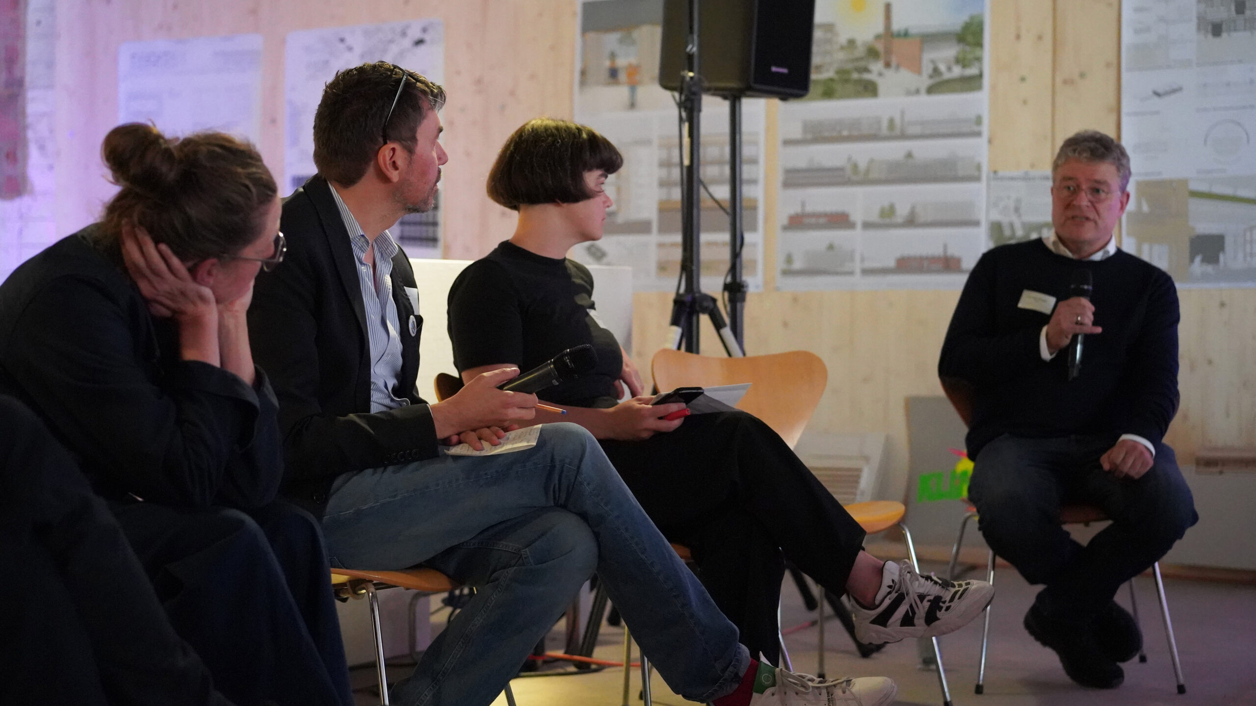 Vier Personen sitzen in einem Innenraum vor Architekturplakaten; drei diskutieren miteinander, während eine Person rechts in ein Mikrofon über transformative Stadtentwicklung spricht. at Hochschule Coburg