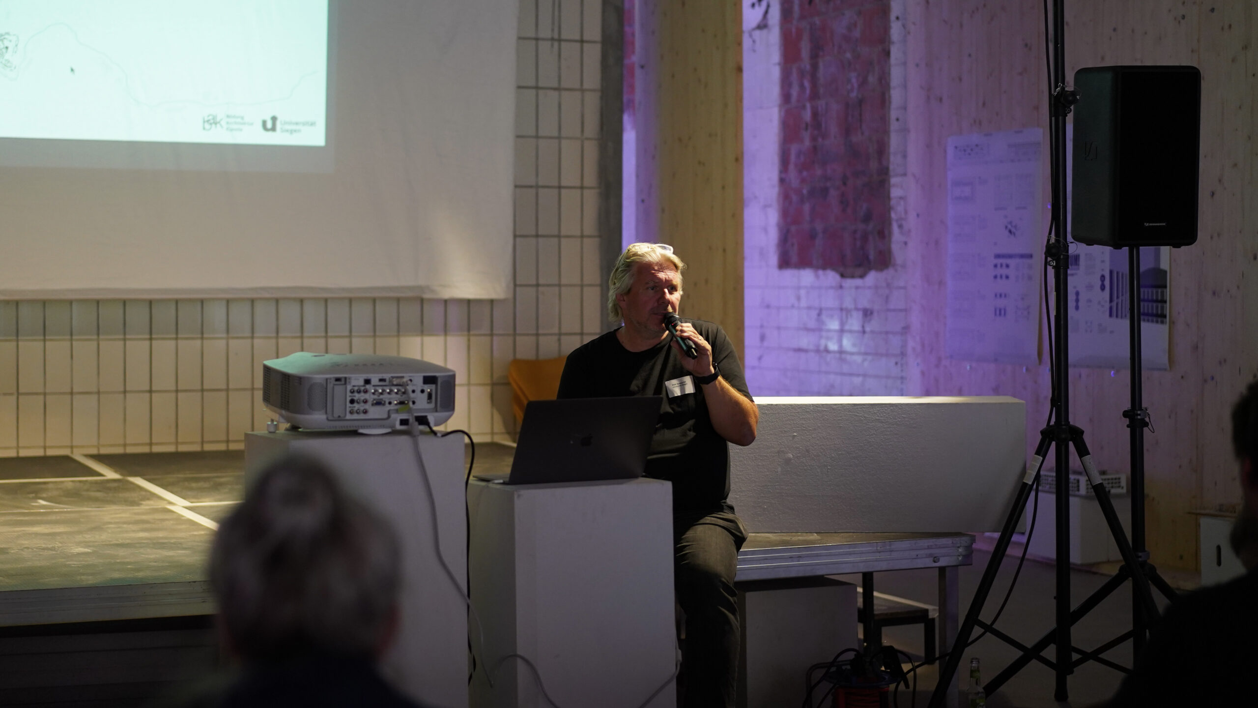 Eine Person mit grauem Haar sitzt in einem schwach beleuchteten Raum neben einem Laptop und einem Projektor und spricht in ein Mikrofon. Auf einer Leinwand hinter ihnen wird eine Präsentation über transformative Stadtentwicklung gezeigt. at Hochschule Coburg