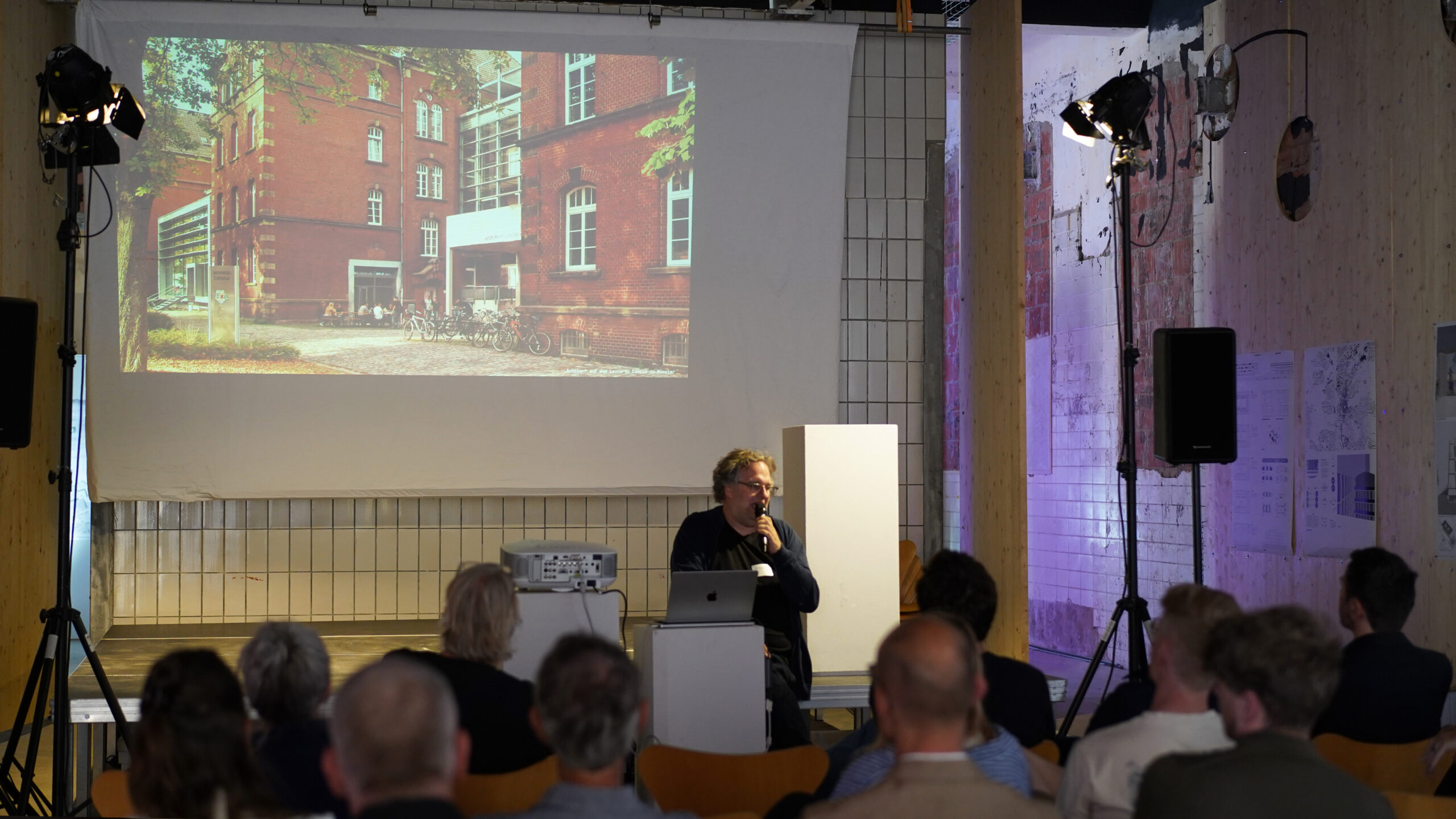 Ein Redner sitzt an einem Podium mit Mikrofon und spricht zu einem Publikum über transformative Stadtentwicklung. Hinter ihm zeigt ein großer Bildschirm das Foto eines roten Backsteingebäudes in einem modernen, gut beleuchteten Raum. at Hochschule Coburg