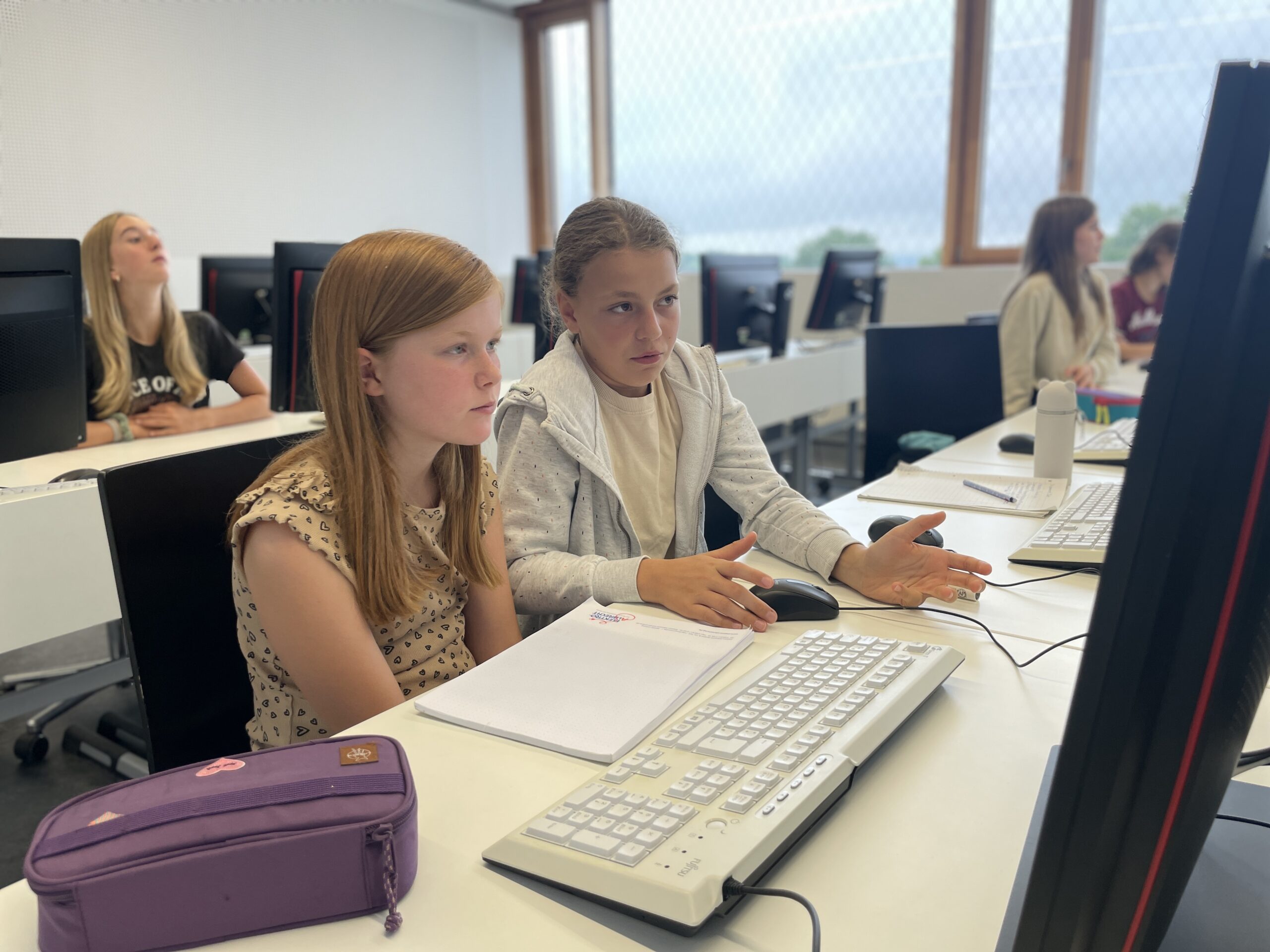 Zwei junge Mädchen sitzen eng zusammen an einem Computer in einem Klassenzimmer und beschäftigen sich mit Technik. Im Hintergrund arbeiten andere Schüler an ihren eigenen Computern. Auf dem Pult liegen ein lila Mäppchen und ein Notizbuch. at Hochschule Coburg