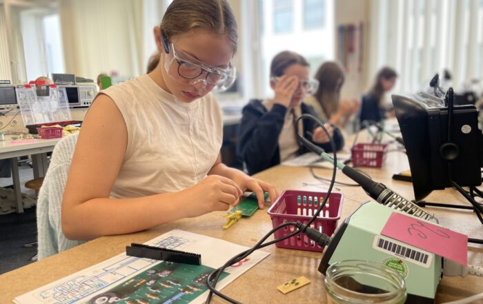 Ein Mädchen, das eine Schutzbrille trägt, arbeitet in einem Klassenzimmer an einem Technik-Projekt mit kleinen Werkzeugen und Platinen. In diesen spannenden Ferien sind auch andere Schüler an einem Tisch mit ähnlichen Aktivitäten beschäftigt. at Hochschule Coburg