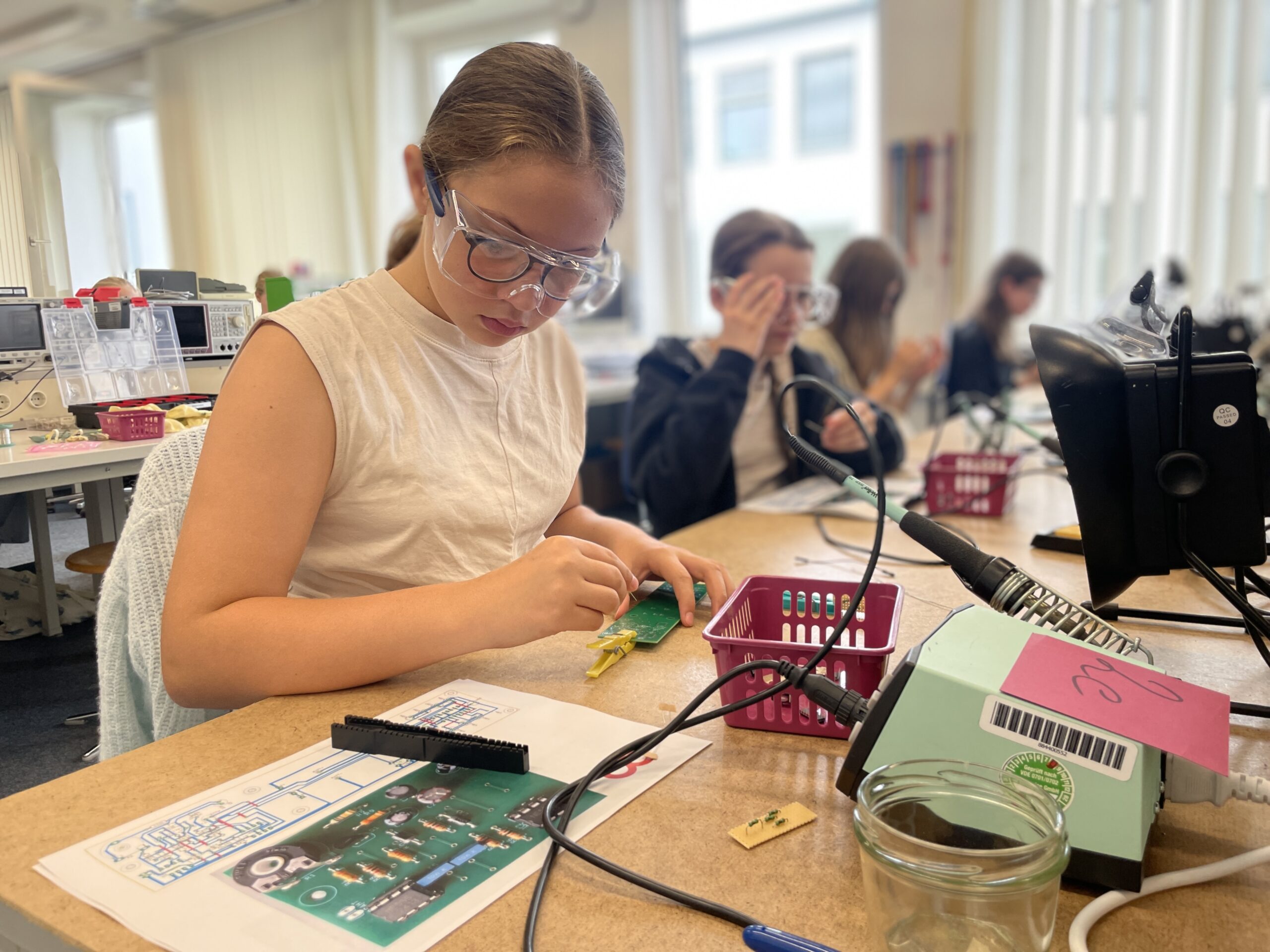 Ein Mädchen, das eine Schutzbrille trägt, arbeitet in einem Klassenzimmer an einem Technik-Projekt mit kleinen Werkzeugen und Platinen. In diesen spannenden Ferien sind auch andere Schüler an einem Tisch mit ähnlichen Aktivitäten beschäftigt. at Hochschule Coburg