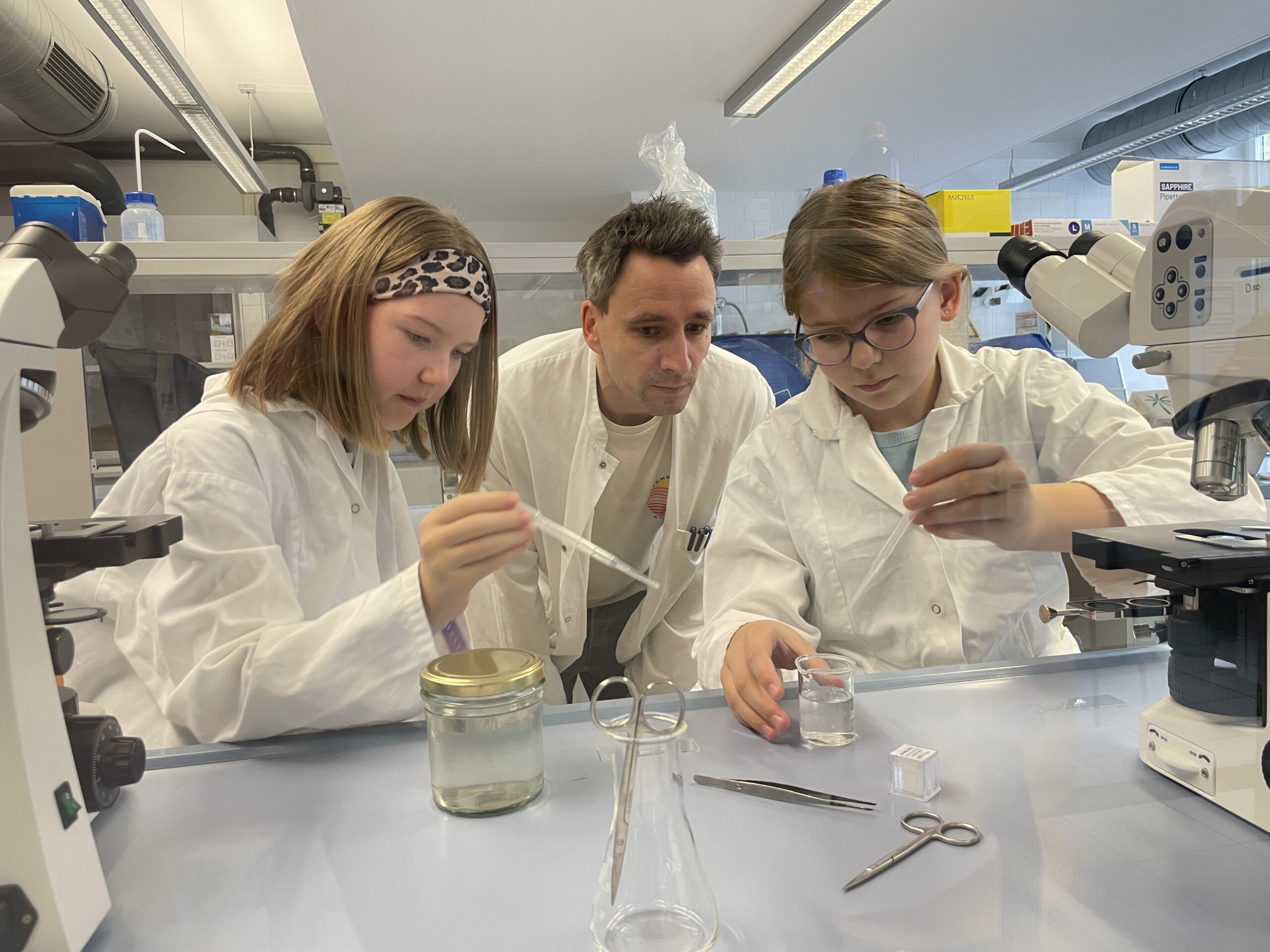 Drei Personen in Laborkitteln arbeiten gemeinsam in einem Labor mit Pipetten und Glasgeräten. Umgeben von Mikroskopen und wissenschaftlichen Geräten konzentrieren sie sich auf ein MUT-Experiment, bei dem es um präzise Technik und Teamarbeit geht. at Hochschule Coburg
