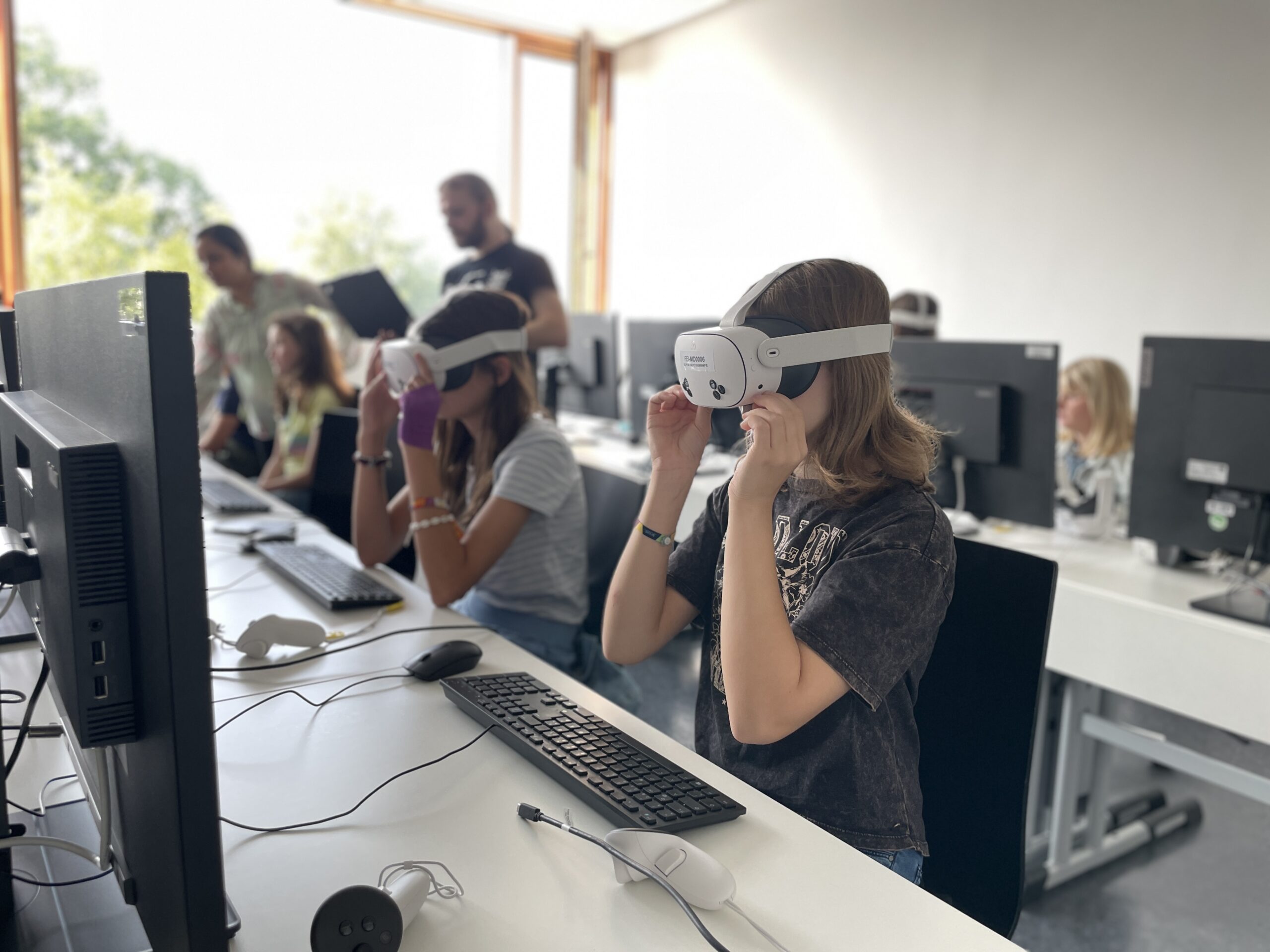 Schüler in einem Klassenzimmer sitzen mit VR-Headsets an Computerschreibtischen und beschäftigen sich mit moderner Technik. Andere Schüler und ein Lehrer sind im Hintergrund zu sehen - einige beobachten, andere nehmen teil - während das Sonnenlicht durch offene Fenster einfällt. at Hochschule Coburg