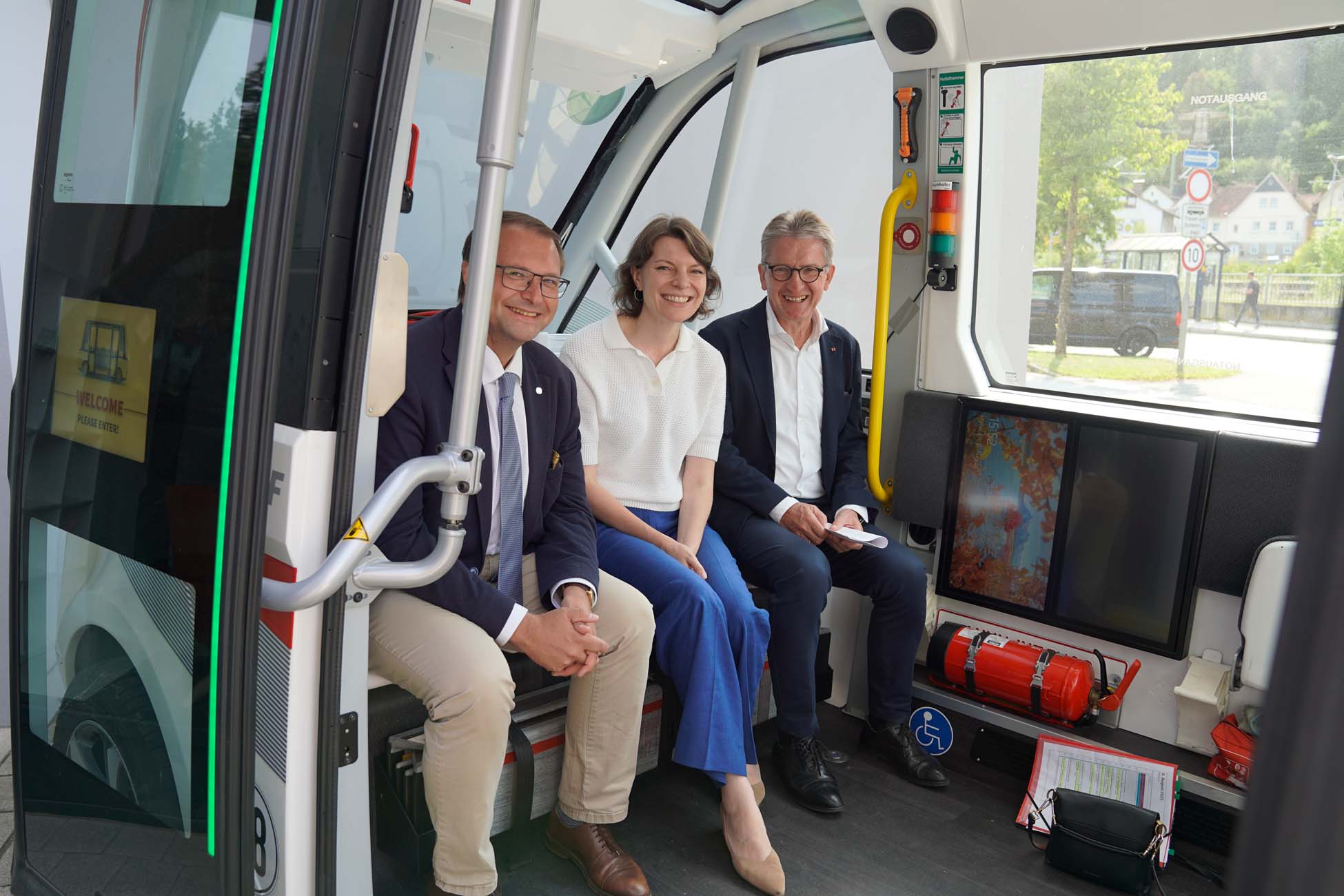 Drei Fachleute in Business-Kleidung sitzen und lächeln in einem modernen, fahrerlosen MoVe-Fahrzeug des öffentlichen Nahverkehrs. Sicherheitseinrichtungen und Hinweisschilder sind zu sehen, durch die Fenster blickt man auf "Bewegung auf dem Land". at Hochschule Coburg