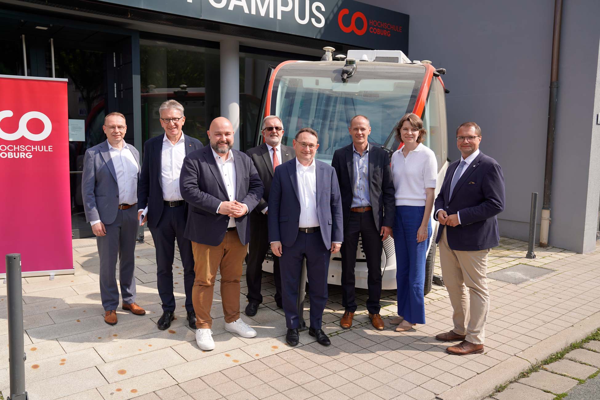 Acht Personen in Business-Kleidung stehen lächelnd vor einem autonomen MoVe-Shuttle-Fahrzeug vor einem Gebäude mit "CAMPUS"-Beschriftung. Links ein rosa "HOCHSCHULE COBURG"-Banner, das auf die Bewegung auf dem Land und den Austausch zwischen Fachleuten und Politik hinweist. at Hochschule Coburg