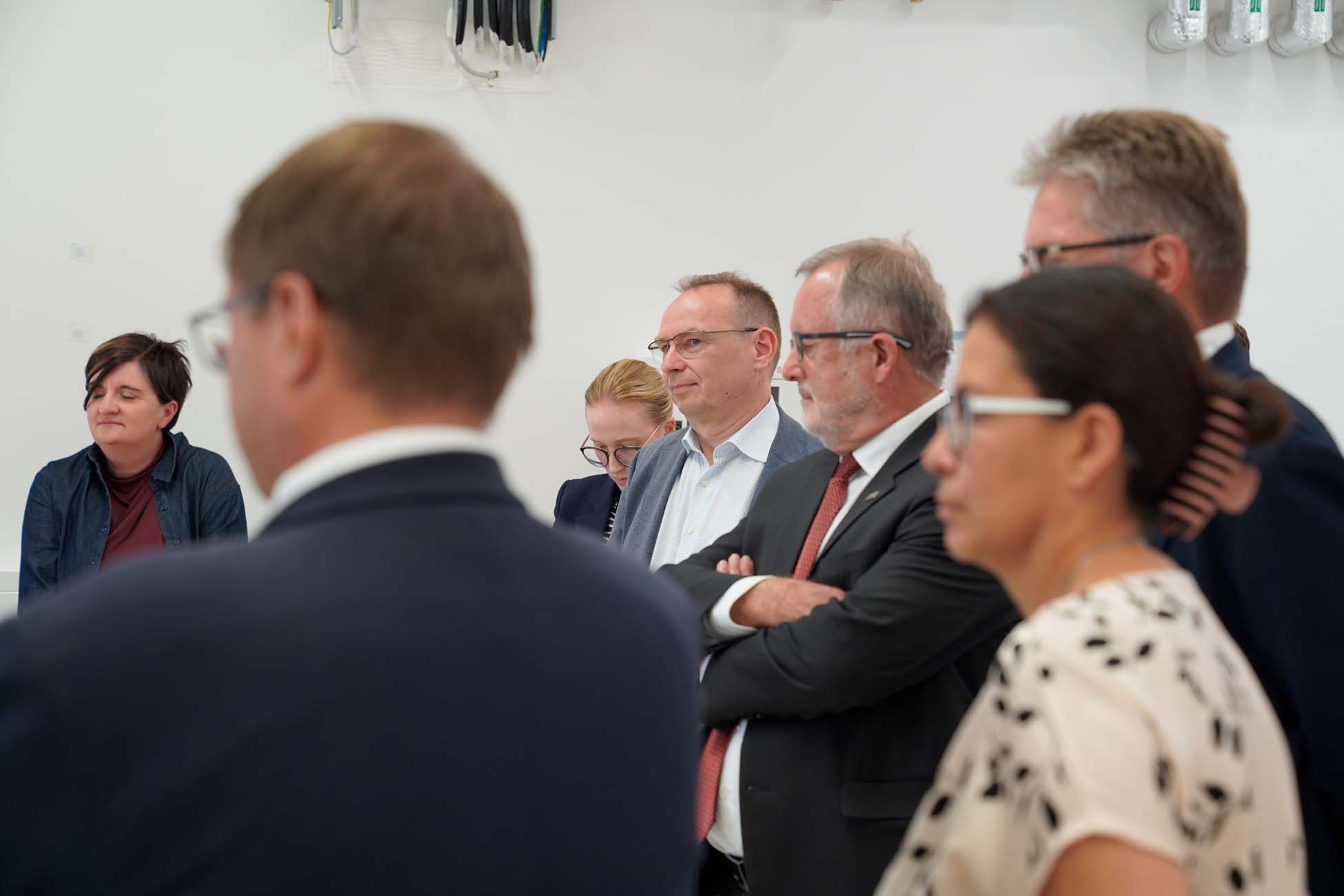 Eine Gruppe von Fachleuten, Männer und Frauen, steht in einem Innenraum zusammen und hört aufmerksam jemandem zu, der nicht zu sehen ist. Die schlichte weiße Wand deutet auf ein Treffen der MoVe oder der Bewegung auf dem Land hin, bei dem der fachliche Austausch im Vordergrund steht. at Hochschule Coburg