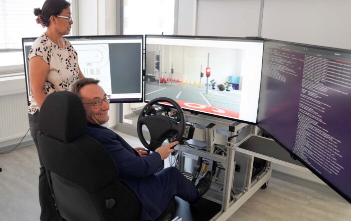 Ein Mann sitzt in einem Fahrsimulator mit drei großen Monitoren, die eine virtuelle Umgebung zeigen, während eine Frau neben ihm steht und beobachtet. Die moderne Laborumgebung erinnert an ein MoVe-Projekt, bei dem Fachleute und Politik innovative Lösungen erproben. at Hochschule Coburg