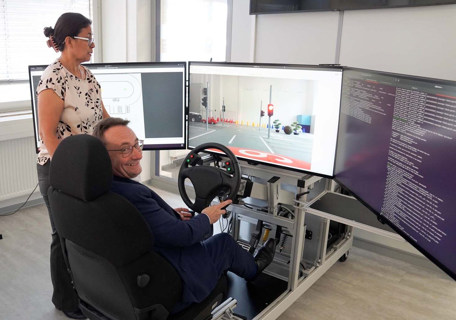 Ein Mann sitzt in einem Fahrsimulator mit drei großen Monitoren, die eine virtuelle Umgebung zeigen, während eine Frau neben ihm steht und beobachtet. Die moderne Laborumgebung erinnert an ein MoVe-Projekt, bei dem Fachleute und Politik innovative Lösungen erproben. at Hochschule Coburg