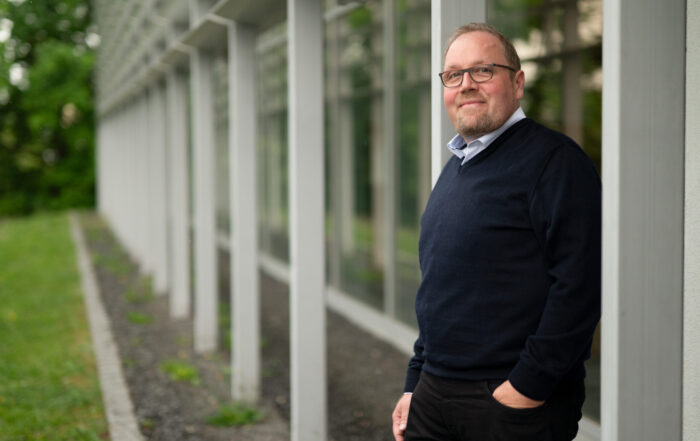 Ein Mann mit Brille und Bart, bekleidet mit einem marineblauen Kanzlerpulli und einer schwarzen Hose, steht im Freien an ein modernes Glasgebäude gelehnt, die Hand in der Tasche, und lächelt in die Kamera. Im Hintergrund ist eine Grünanlage zu sehen. at Hochschule Coburg