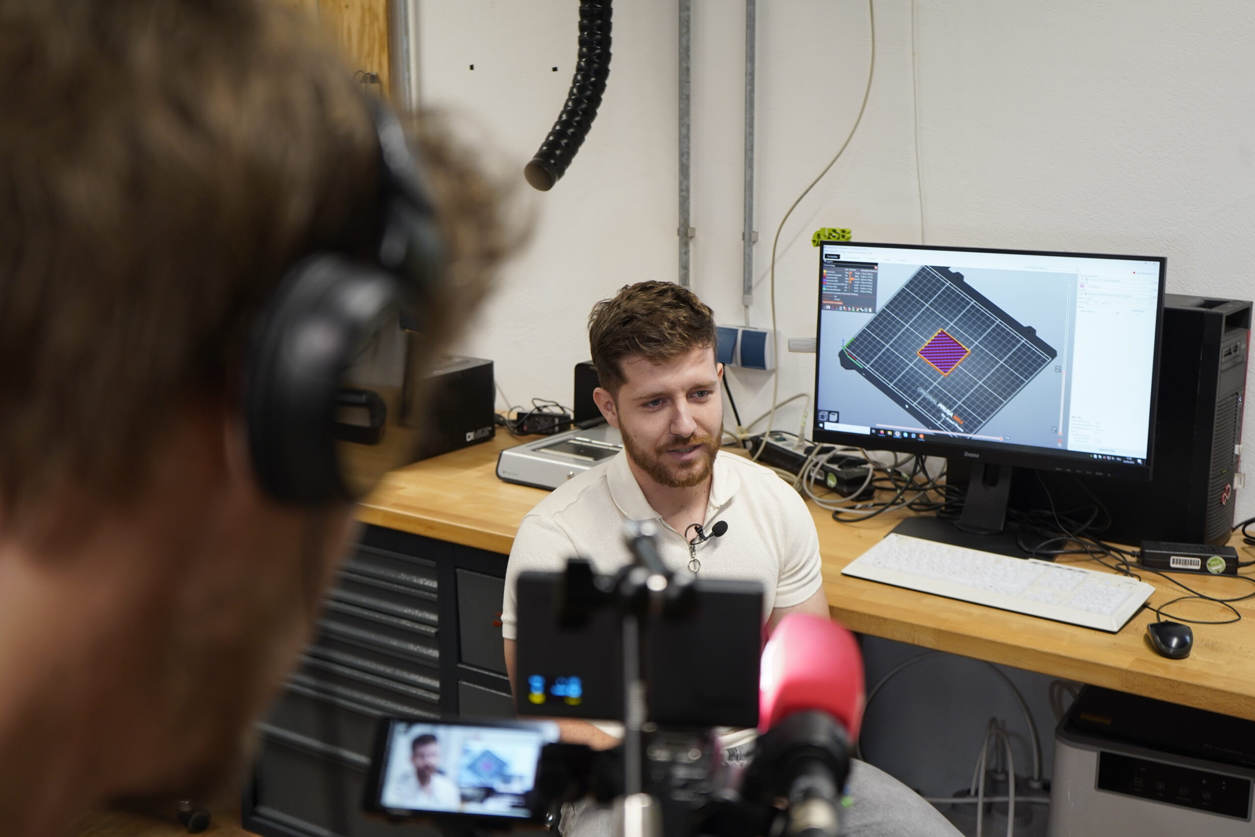 Ein Mann sitzt vor einem Computer, der ein 3D-Modell anzeigt, und wird von der Kamera in einer innovativen Hochschul-Umfeld-Werkstatt interviewt. Auf der Werkbank sind Geräte und Werkzeuge zu sehen. at Hochschule Coburg