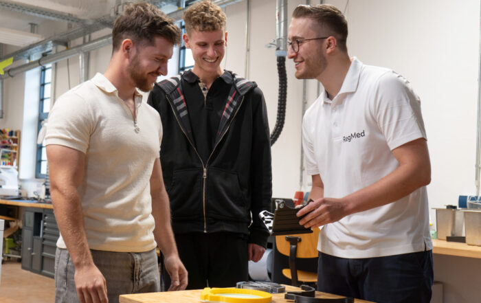 Drei junge Männer stehen in einer Werkstatt um einen Tisch herum und lächeln über einen schwarzen Gegenstand, den einer von ihnen in der Hand hält. Umgeben von Werkzeugen und Geräten diskutieren sie in dieser kreativen Umgebung über Innovation Hochschul-Umfeld und Ideen für Pferde Therapie. at Hochschule Coburg
