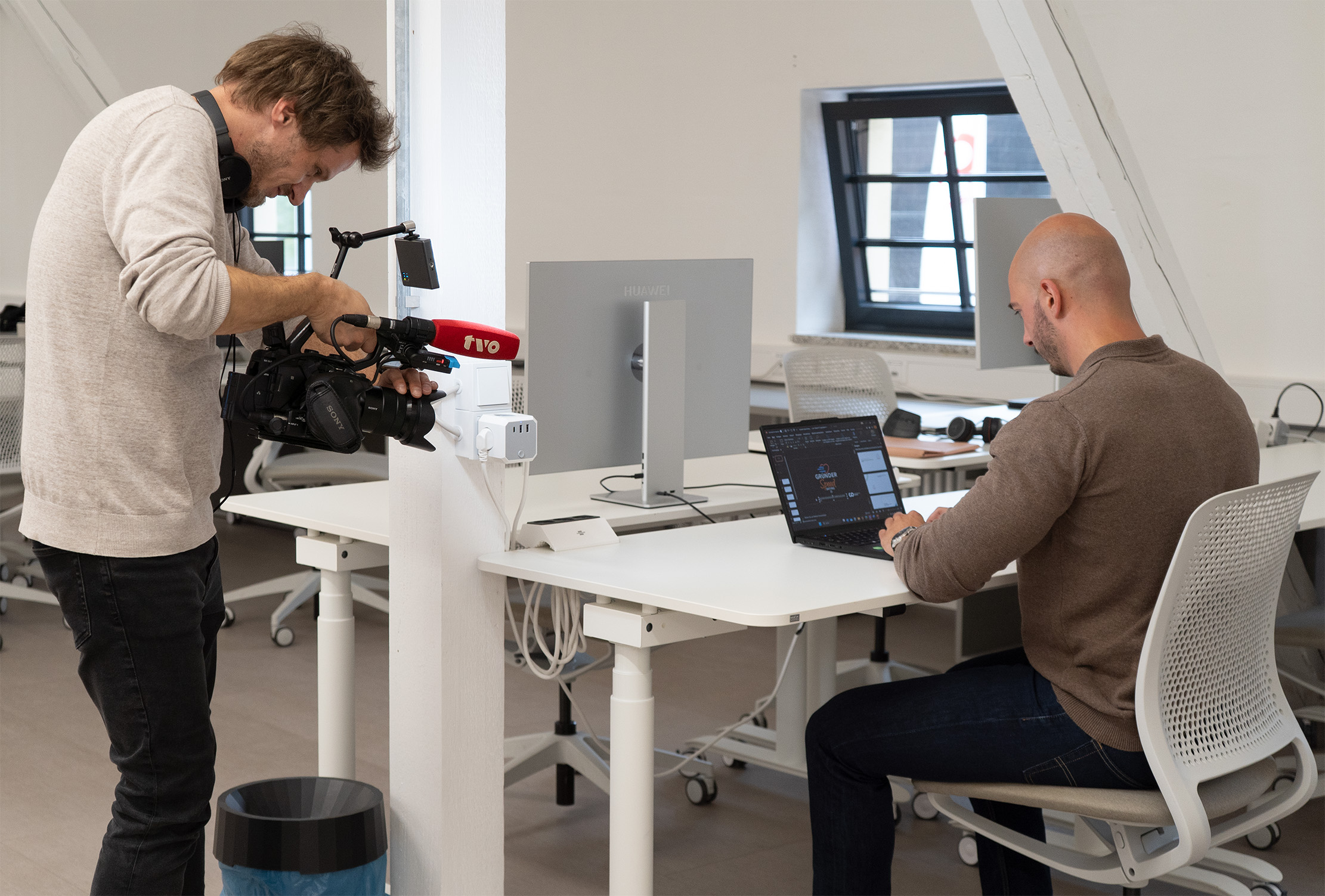 Eine Person sitzt an einem Schreibtisch und arbeitet an einem Laptop, während eine andere Person in der Nähe steht und eine Videokamera mit der Aufschrift "TV2" auf die sitzende Person in einem modernen Büroraum richtet und ein Interview über Innovation in der Stammzelltherapie für Pferde aufnimmt. at Hochschule Coburg