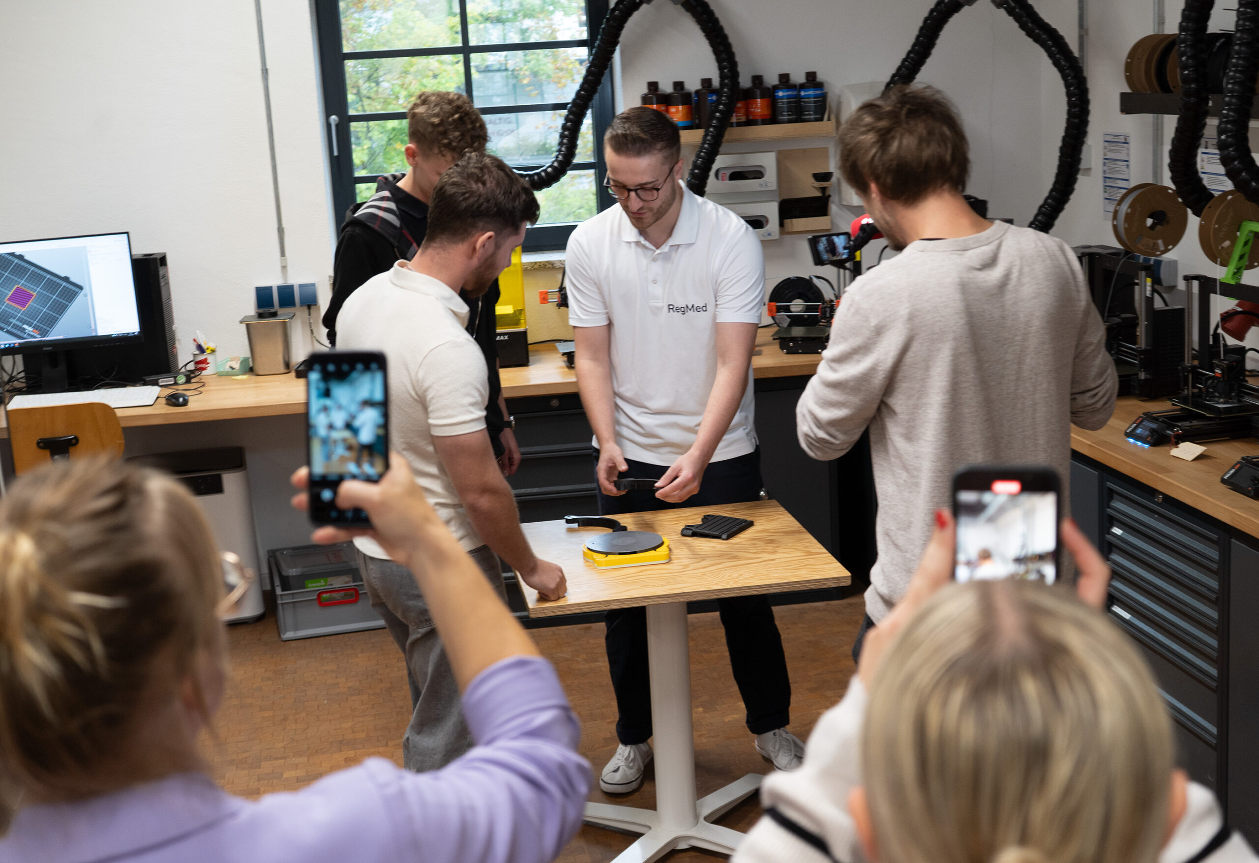 In einem Hochschul-Umfeld versammelt sich eine Gruppe um einen Tisch in einer Werkstatt und sieht zu, wie ein Mann in einem weißen Hemd ein innovatives Gerät vorführt; zwei Personen im Vordergrund machen Fotos oder Videos mit ihren Smartphones. at Hochschule Coburg