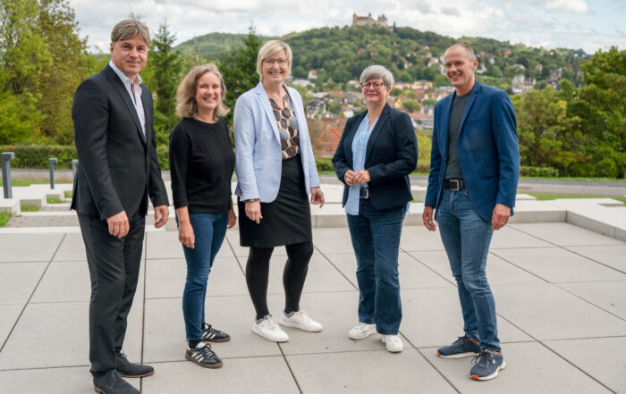 Fünf Erwachsene - drei Frauen und zwei Männer - stehen im Freien auf einer gepflasterten Fläche und lächeln in die Kamera. Hinter ihnen sind Bäume, ein Hügel und ein Schloss unter einem teilweise bewölkten Himmel zu sehen, was die Zusammenarbeit zwischen der Hochschule Coburg und den Schulen Südthüringen unterstreicht. at Hochschule Coburg