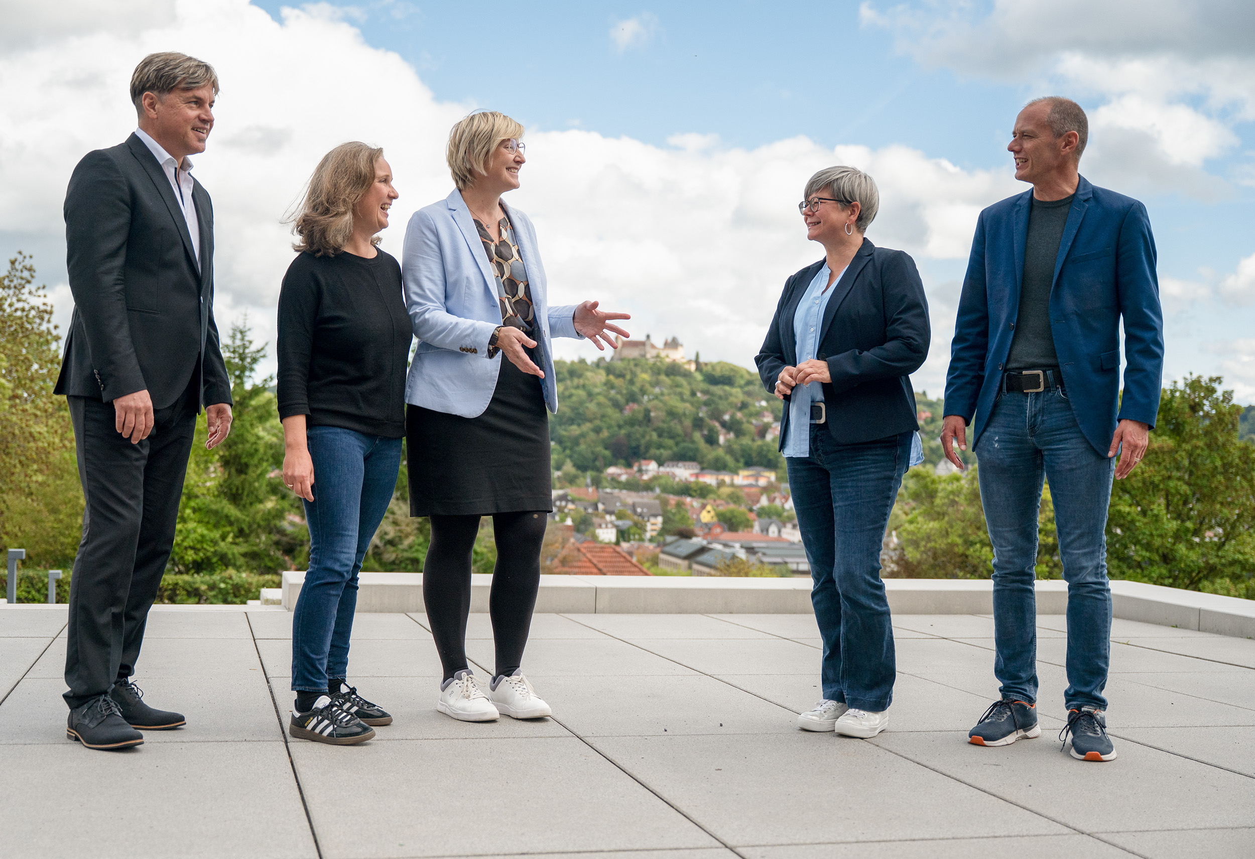 Fünf Erwachsene stehen im Freien auf einer Terrasse und sprechen und lächeln über Zusammenarbeit. Im Hintergrund sind grüne Hügel und eine Stadt in Südthüringen zu sehen, der Himmel ist teilweise bewölkt. at Hochschule Coburg