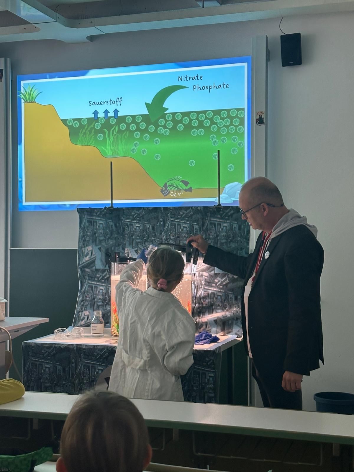 Ein Kind in einem Laborkittel und ein Erwachsener, möglicherweise Professorinnen und Professoren, führen vor einer Klasse ein wissenschaftliches Experiment vor. Hinter ihnen zeigt ein großer Bildschirm ein Diagramm über Sauerstoff, Nitrat und Phosphat in Boden und Wasser. at Hochschule Coburg