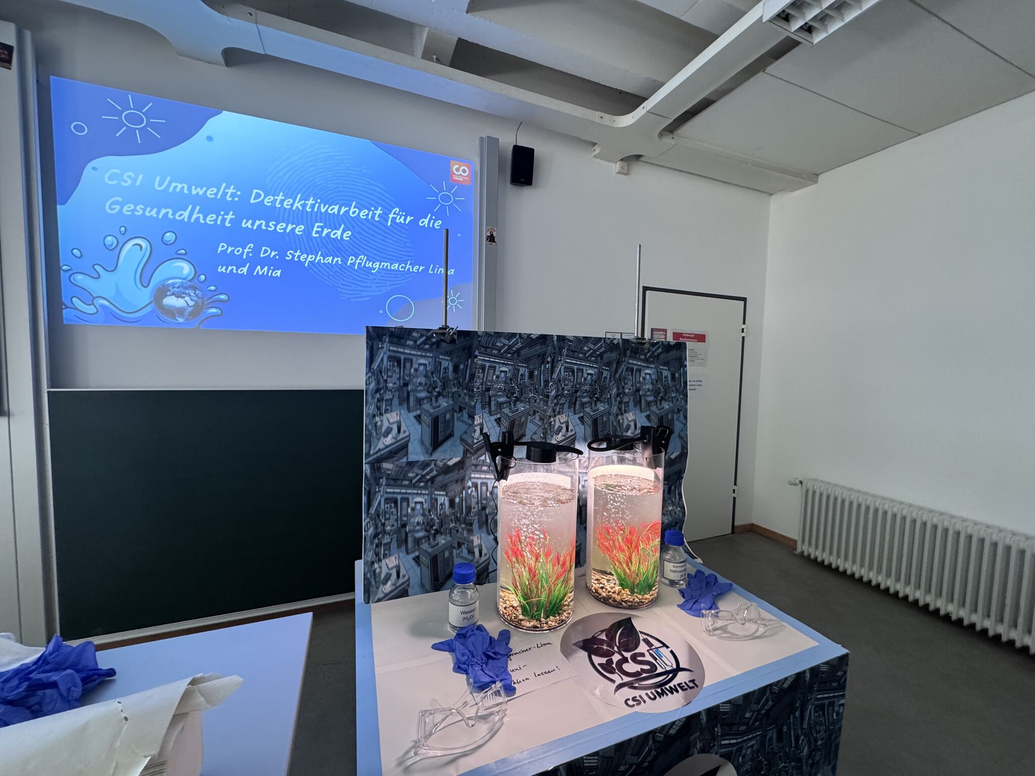 Ein Klassenzimmer mit einer Projektionsfläche, auf der "CSI Umwelt: Detektivarbeit für die Gesundheit unserer Erde". Professorinnen und Professoren inspirieren Kinder, indem sie Wasserpflanzen, Laborgloves und Schutzbrillen auf dem Tisch präsentieren. at Hochschule Coburg