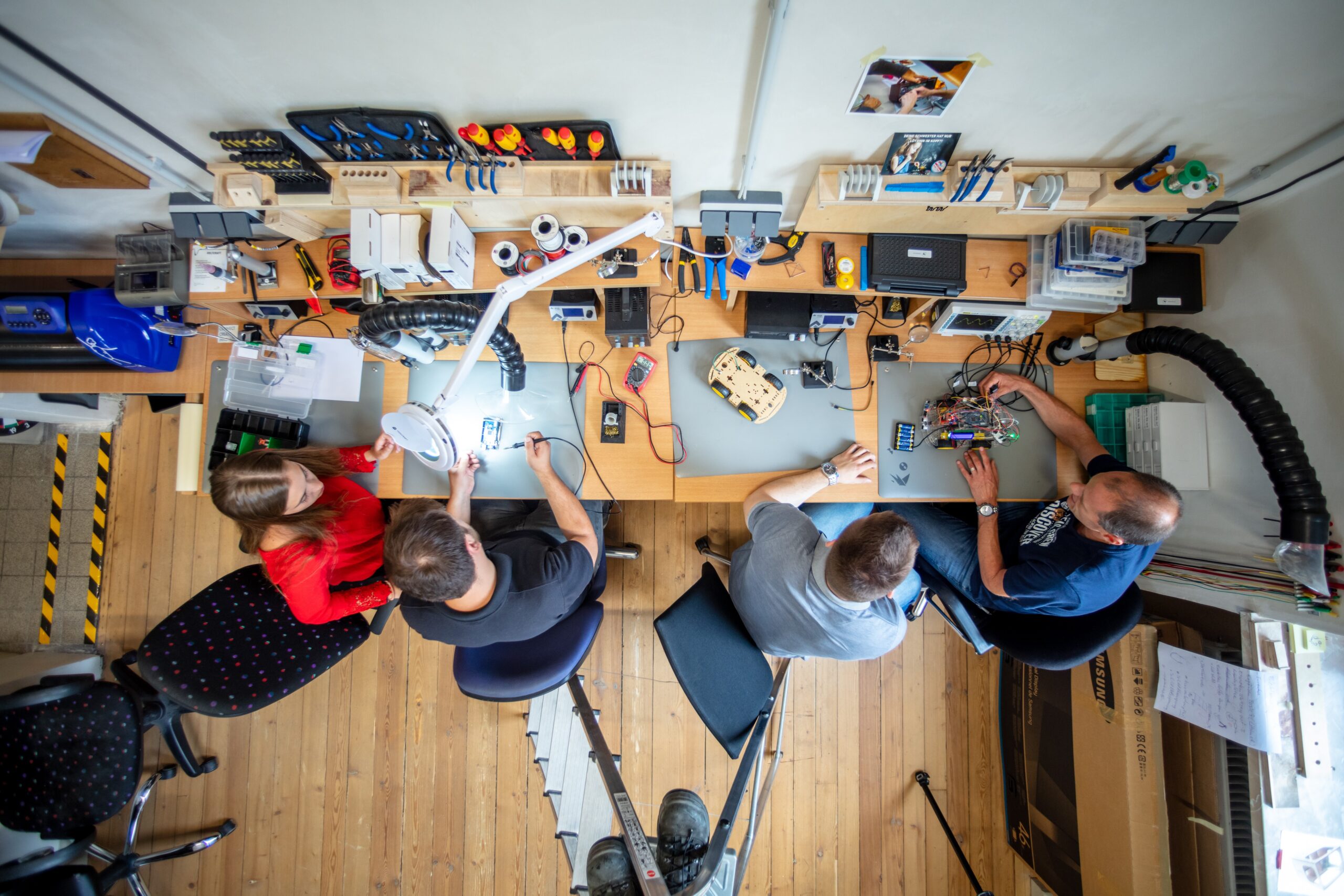 Vier Personen sitzen an einer Werkbank, von oben gesehen, und arbeiten mit elektronischen Bauteilen und Werkzeugen in einem hellen, organisierten Labor mit Holzboden und verschiedenen Geräten auf den Tischen. at Hochschule Coburg