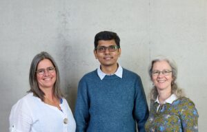 Drei Erwachsene stehen Seite an Seite vor einer schlichten hellen Wand und lächeln in die Kamera. Die beiden Frauen tragen Brillen und lässige Oberteile, während der Mann in der Mitte, bekleidet mit einem blauen Pullover, Begeisterung für Informatik und Technik mitbringt. at Hochschule Coburg