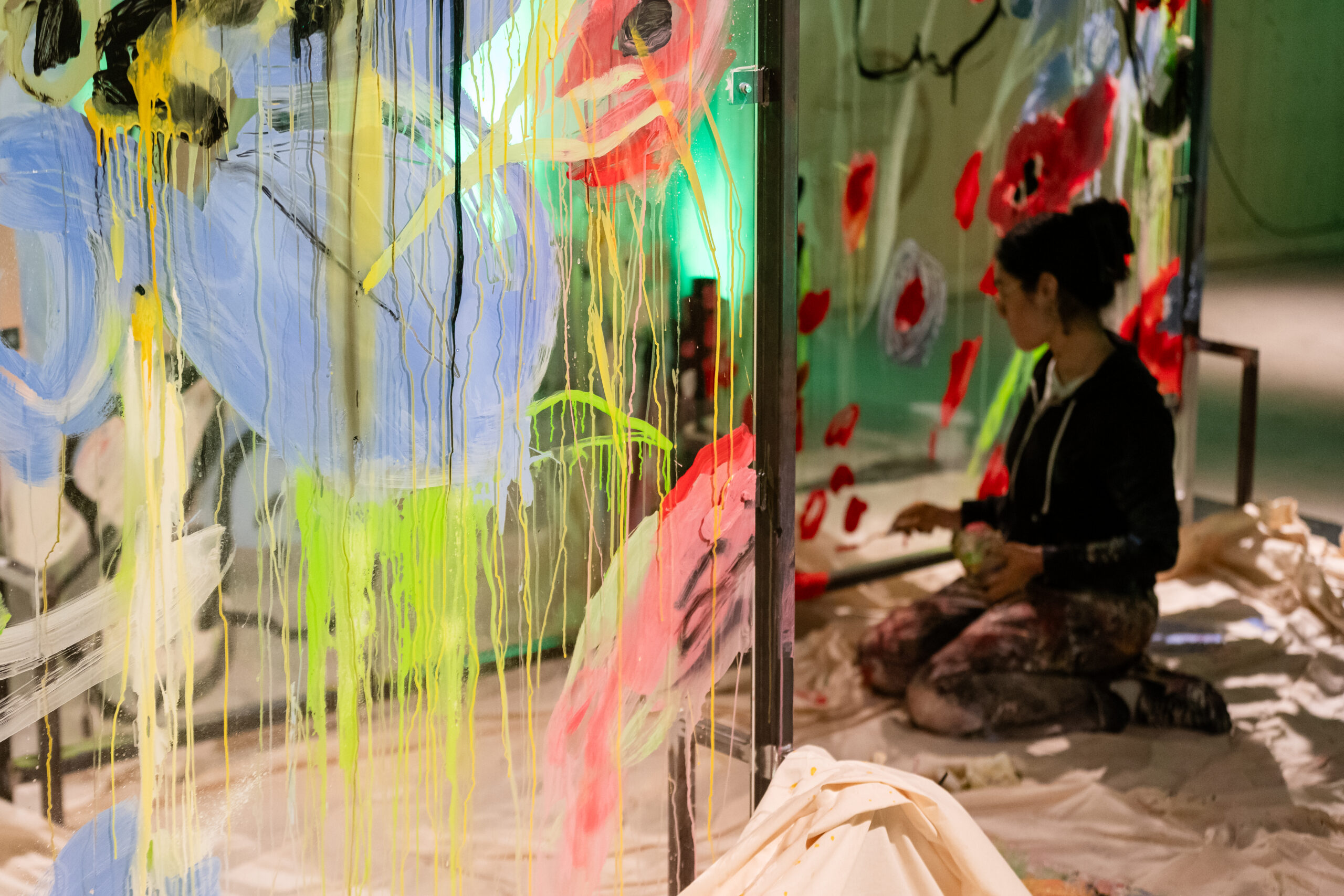 Eine Person sitzt auf dem Boden und malt mit blauer, gelber und roter Farbe lebhafte abstrakte Formen und Linien auf eine transparente Platte in einem Kunstatelier, umgeben von Stoffen und Kunstzubehör. at Hochschule Coburg