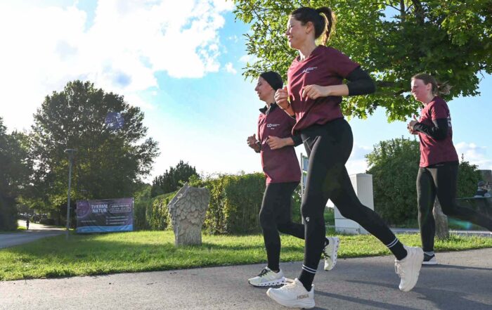 Drei Personen in kastanienbraunen Hemden und schwarzen Leggings joggen an einem sonnigen Tag im Freien, wahrscheinlich bei einem Firmenlauf in der Nähe einer Hochschule, mit Bäumen und Grünflächen im Hintergrund. Die Sonne scheint hell von der linken Seite des Bildes. at Hochschule Coburg