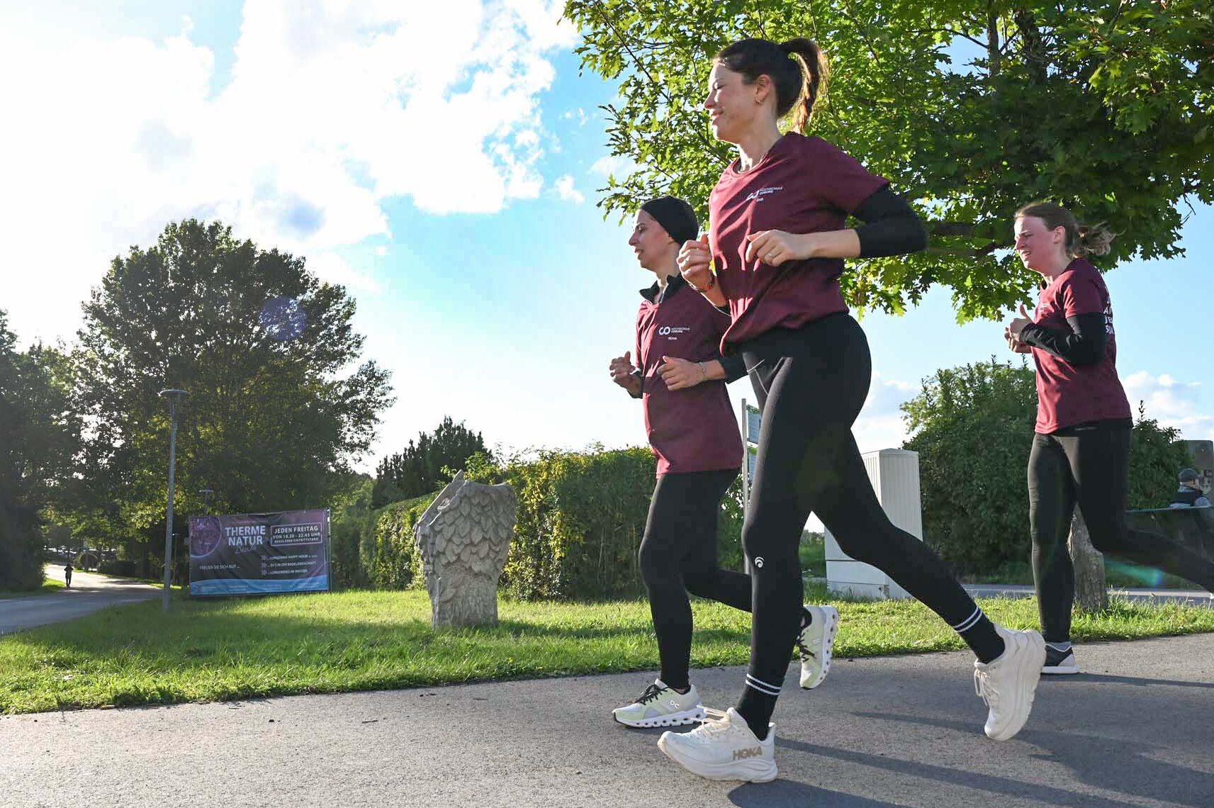 Drei Personen in kastanienbraunen Hemden und schwarzen Leggings joggen an einem sonnigen Tag im Freien, wahrscheinlich bei einem Firmenlauf in der Nähe einer Hochschule, mit Bäumen und Grünflächen im Hintergrund. Die Sonne scheint hell von der linken Seite des Bildes. at Hochschule Coburg