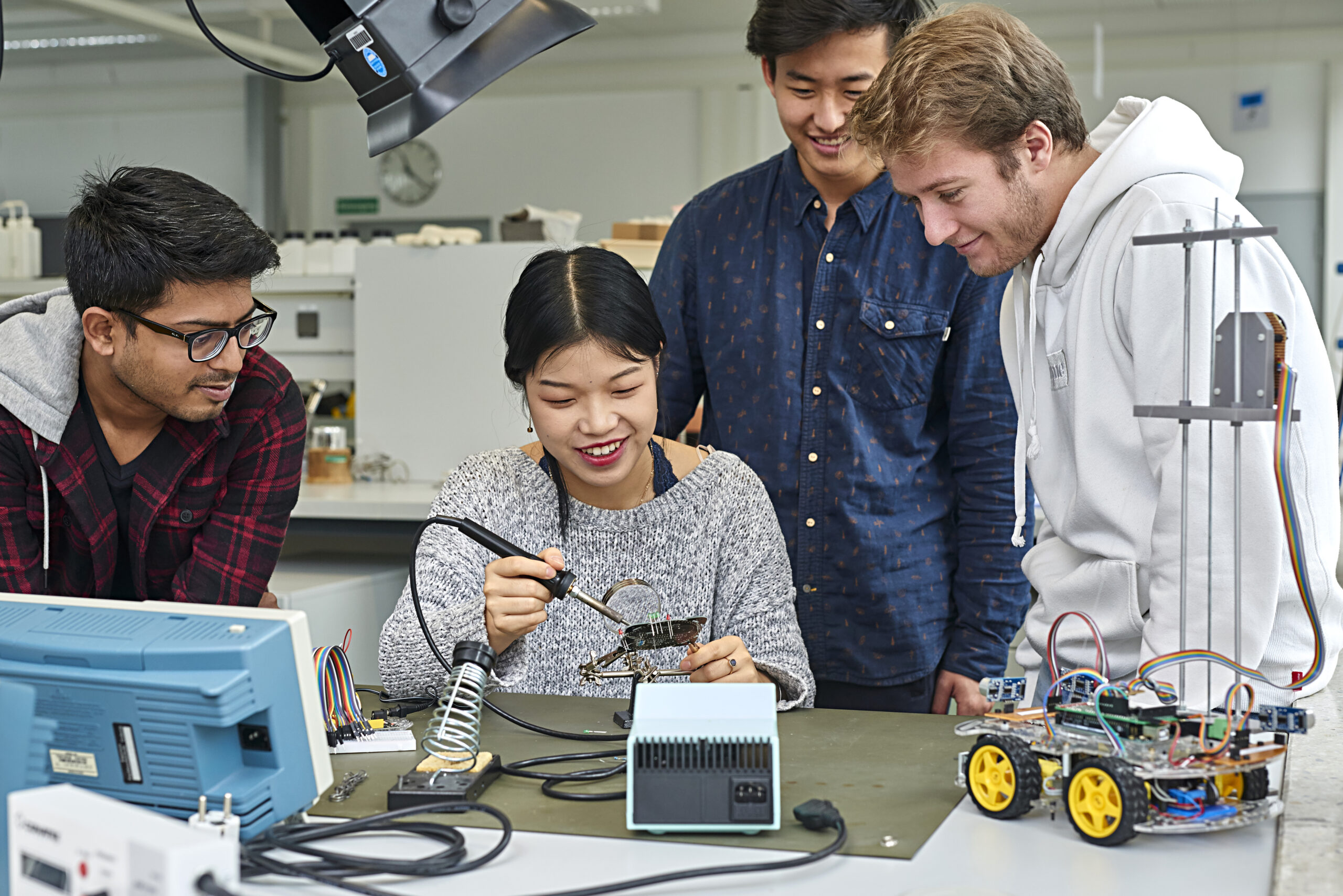 Vier junge Erwachsene versammeln sich um einen Schreibtisch in einem Labor und sehen zu, wie eine Person mit einem Lötkolben an Elektronik arbeitet, umgeben von Leiterplatten und einem kleinen Roboter. at Hochschule Coburg