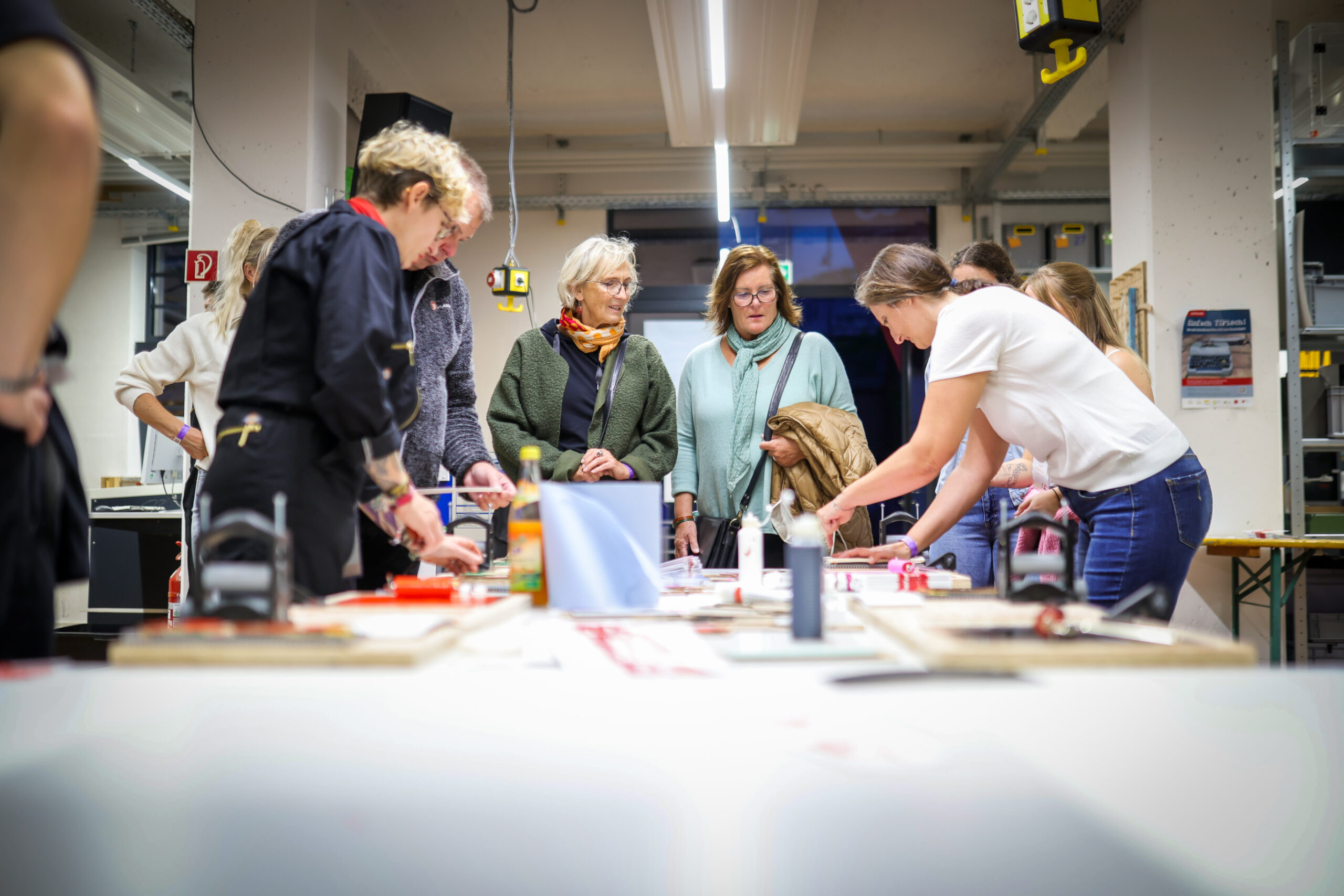 Eine Gruppe von Menschen versammelt sich in einer hellen Werkstatt um einen Tisch mit Kunstzubehör und Werkzeugen und konzentriert sich auf ein kreatives Projekt, während eine Person etwas erklärt oder vorführt. at Hochschule Coburg