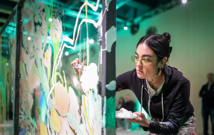 Bei einer Live-Painting-Veranstaltung des Kunstateliers malt Anna Deller-Yee, eine junge Frau mit Brille und schwarzem Kapuzenpulli, konzentriert bunte abstrakte Blumen auf eine große Glasplatte, in der einen Hand eine Palette. at Hochschule Coburg