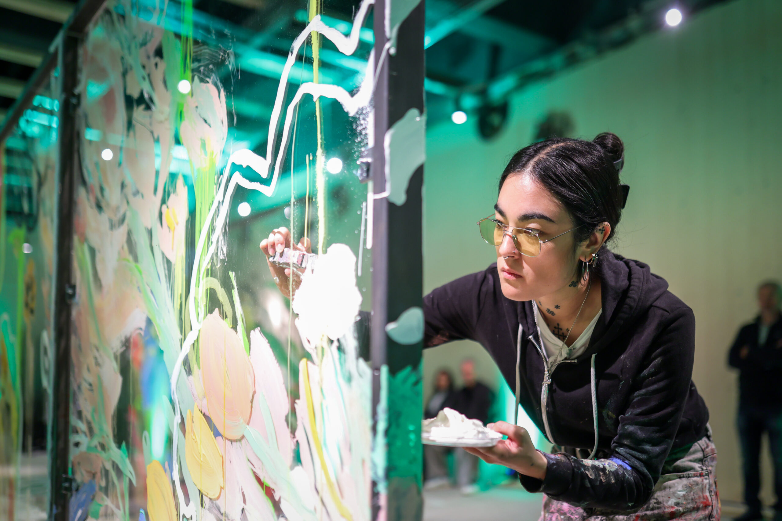Bei einer Live-Painting-Veranstaltung des Kunstateliers malt Anna Deller-Yee, eine junge Frau mit Brille und schwarzem Kapuzenpulli, konzentriert bunte abstrakte Blumen auf eine große Glasplatte, in der einen Hand eine Palette. at Hochschule Coburg