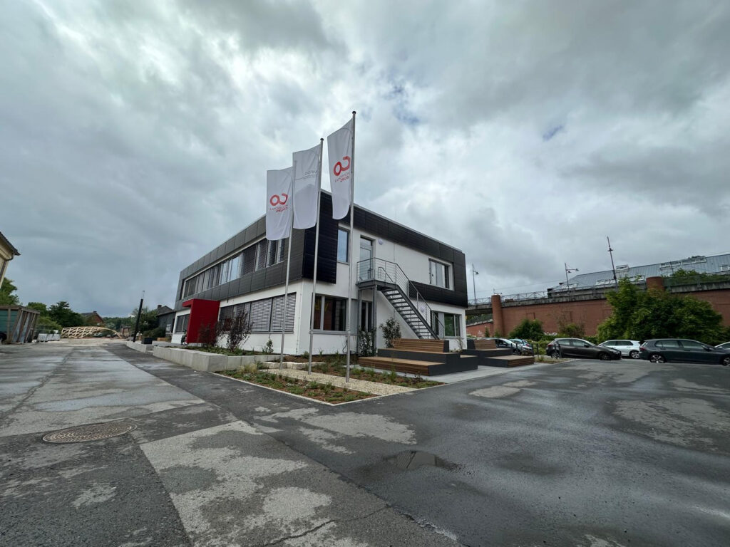 A modern two-story building at Coburg University features three white flags with red logos out front. On this cloudy day, the wet ground reflects several parked cars. Lush plants and a striking red accent wall welcome visitors by the entrance. at Hochschule Coburg