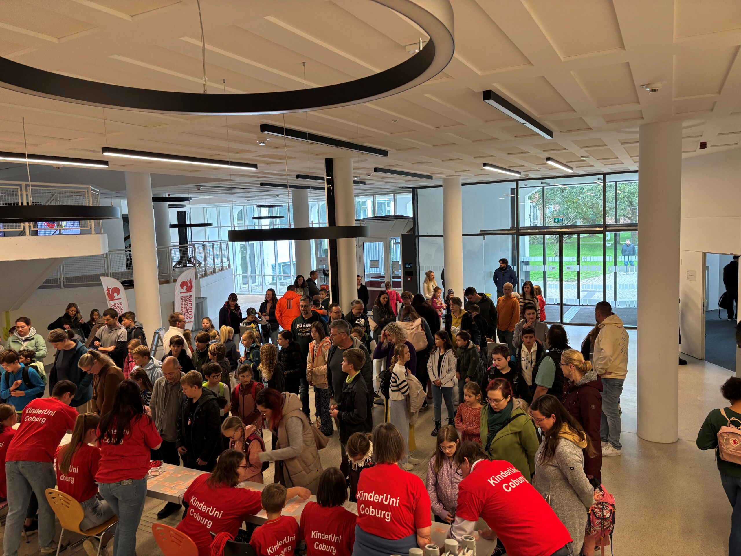Eine große Gruppe von Menschen, darunter viele Kinder und Erwachsene, versammelt sich in einem hellen Gebäude. Mehrere in roten "KinderUni Coburg"-Shirts inspirieren und unterstützen die Besucher am Registrierungs- oder Infotisch wie Professoren. at Hochschule Coburg