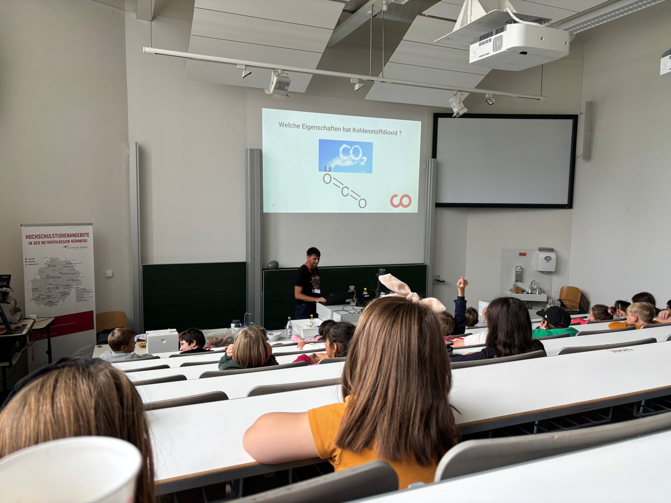 Ein Klassenzimmer mit Kindern, die an Tischen sitzen und einem Professor gegenüberstehen, der an einer Tafel steht und eine Folie über Kohlendioxid projiziert. Einige SchülerInnen heben die Hände, inspiriert und eifrig dabei. at Hochschule Coburg
