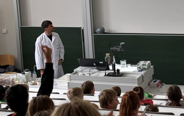 Ein Professor im Laborkittel steht vorne in einem Hörsaal und führt den Kindern eine wissenschaftliche Demonstration vor, während auf der Leinwand ein Reagenzglas-Experiment gezeigt wird. Das Publikum schaut aufmerksam zu, einige heben ihre Hände, offensichtlich inspiriert von der Lektion. at Hochschule Coburg