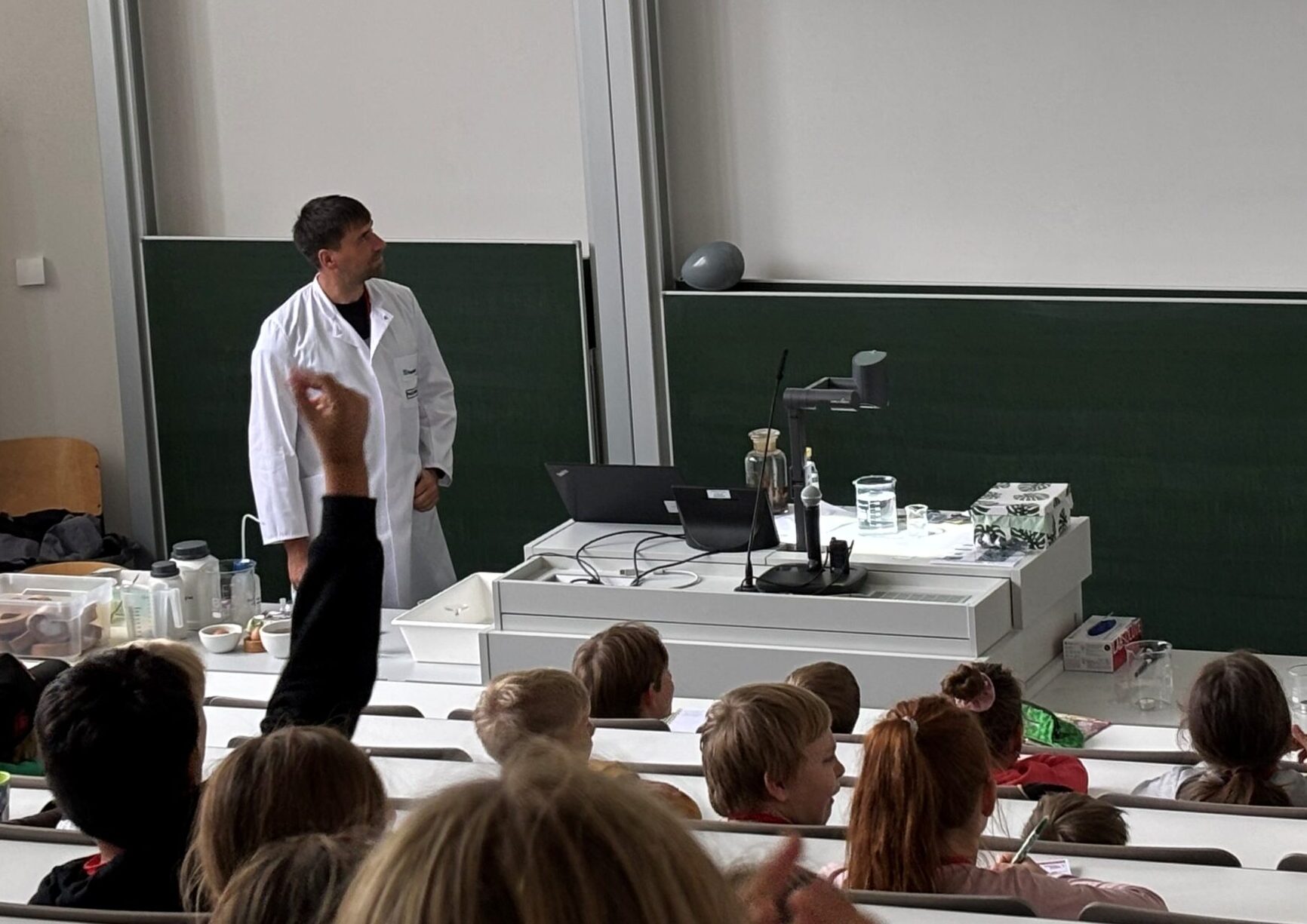 Ein Professor im Laborkittel steht vorne in einem Hörsaal und führt den Kindern eine wissenschaftliche Demonstration vor, während auf der Leinwand ein Reagenzglas-Experiment gezeigt wird. Das Publikum schaut aufmerksam zu, einige heben ihre Hände, offensichtlich inspiriert von der Lektion. at Hochschule Coburg