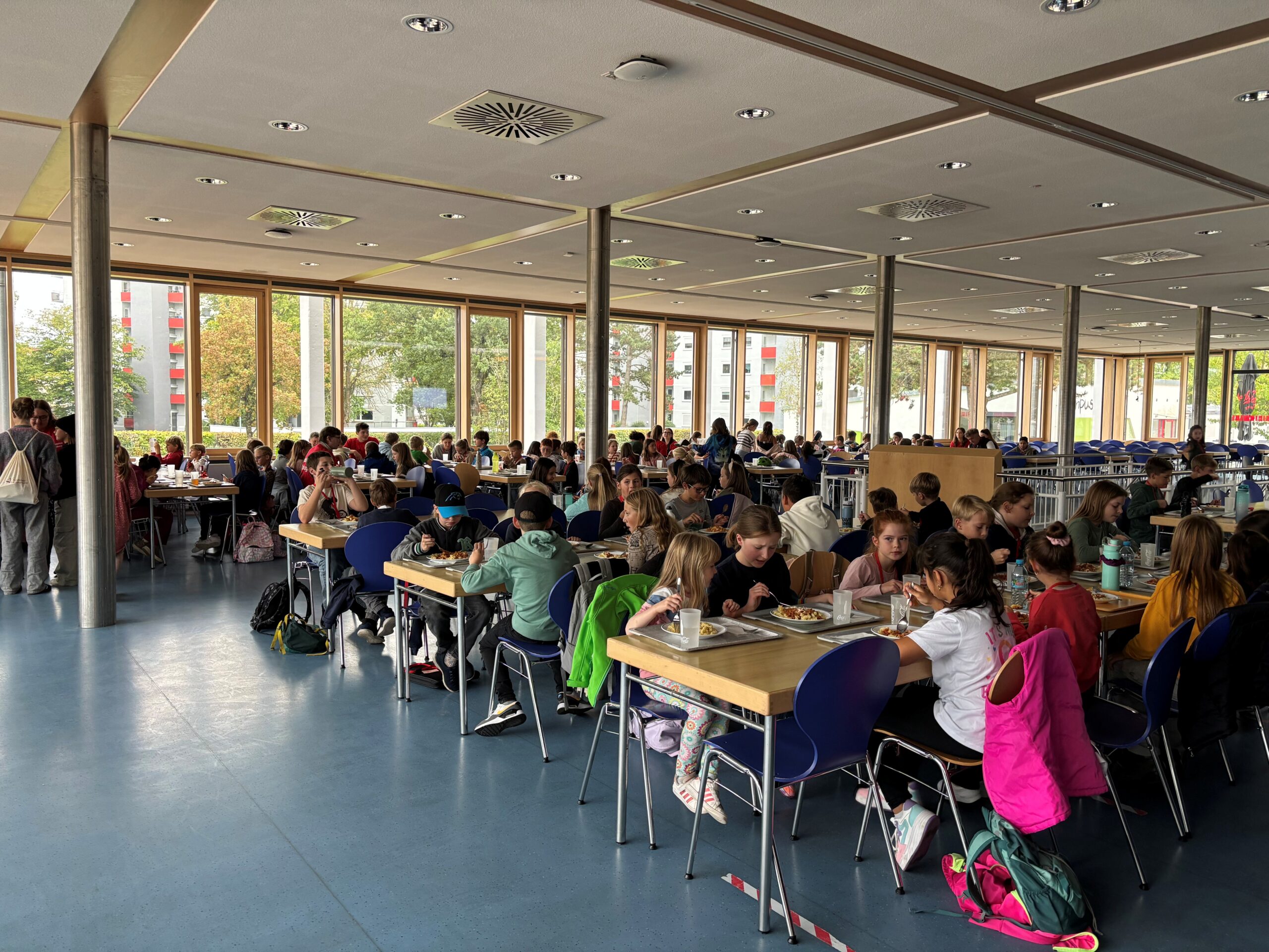Eine große Cafeteria, in der viele Kinder an Tischen sitzen und zu Mittag essen. Der Raum hat große Fenster, die natürliches Licht hereinlassen, und man sieht auch Gruppen von Kindern, die im Hintergrund stehen und sich unterhalten, ihr Lachen inspiriert andere in der Nähe. at Hochschule Coburg