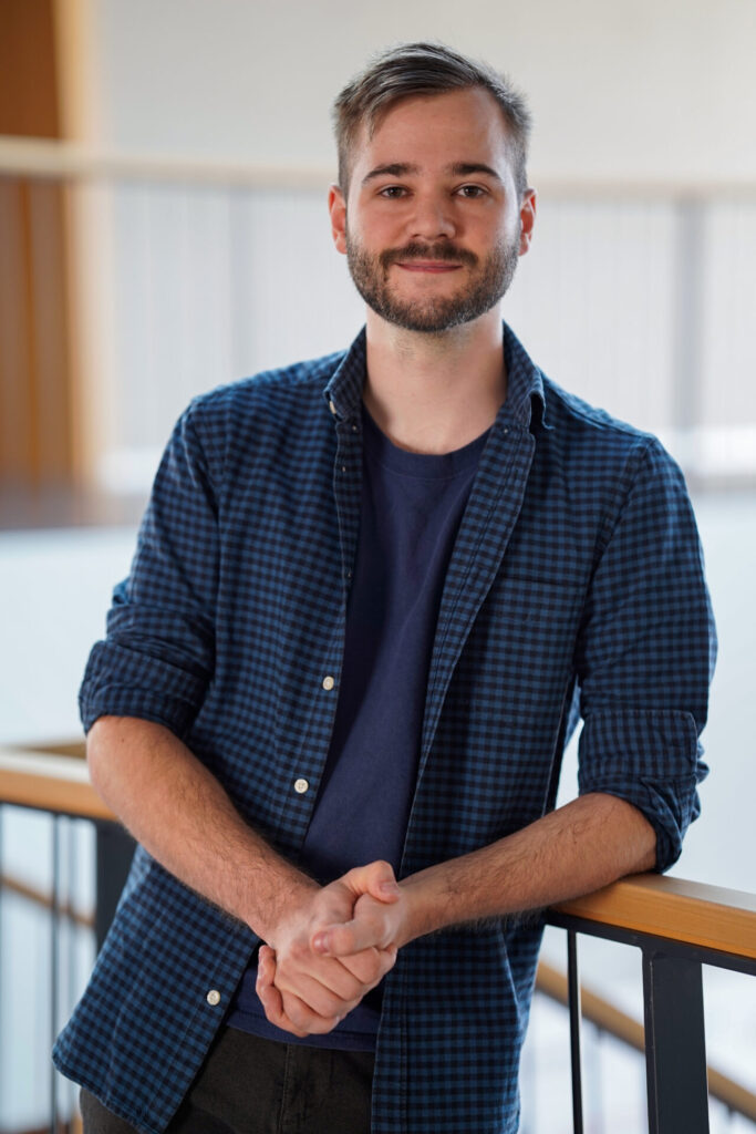 Ein junger Mann mit kurzen Haaren und Bart, der ein blau-kariertes Hemd über einem marineblauen T-Shirt trägt, steht in einem Haus, lehnt sich an ein Geländer und lächelt in die Kamera. at Hochschule Coburg