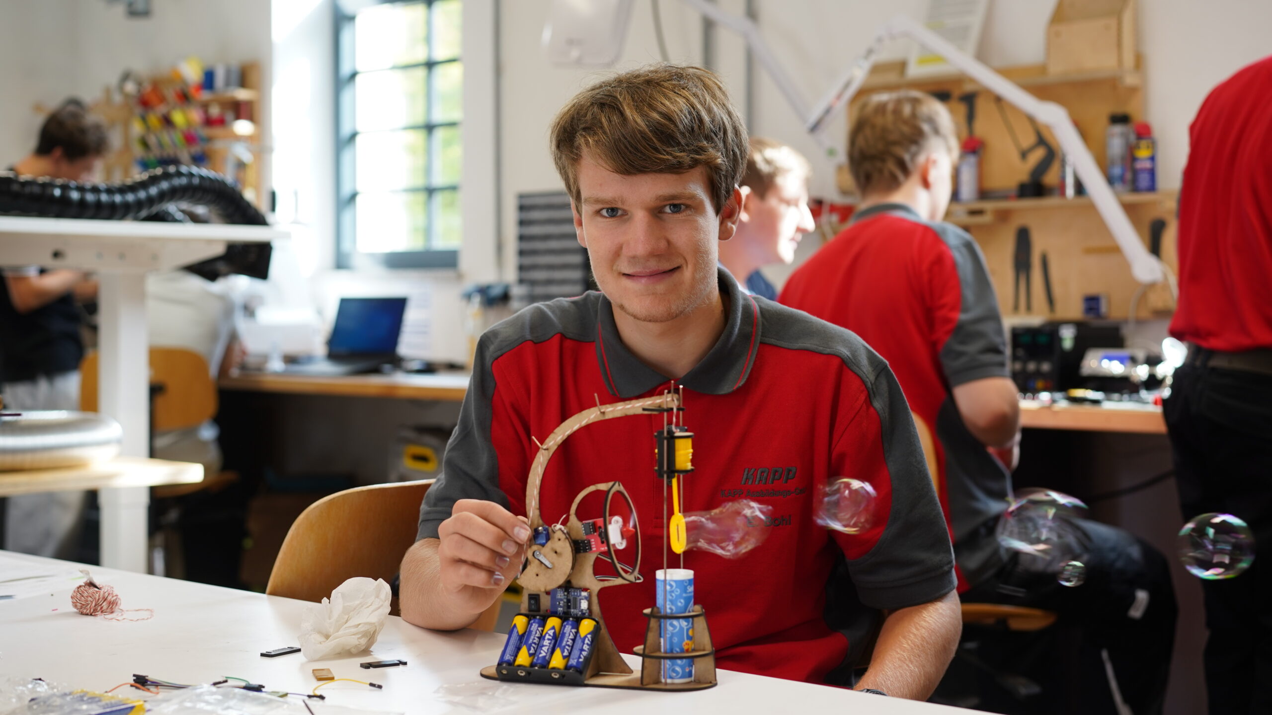 Ein junger Mann in einem rot-grauen Hemd sitzt lächelnd an einem Tisch und hält ein kleines mechanisches Gerät mit Batterien und einem Holzrahmen in der Hand. In der hellen Werkstatt des CREAPOLIS Makerspace blüht der Teamgeist, während im Hintergrund andere zusammenarbeiten. at Hochschule Coburg
