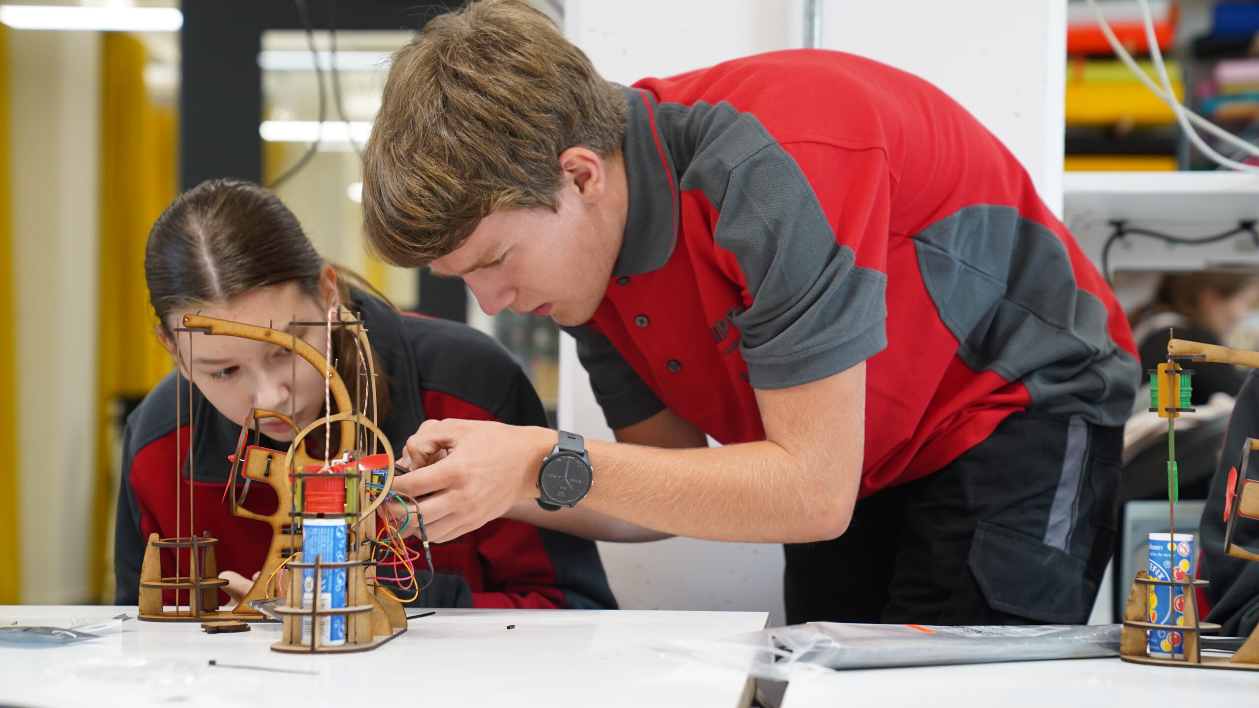 Zwei Schüler in roten und grauen Uniformen demonstrieren Teamgeist, während sie gemeinsam an einem hölzernen Konstruktionsprojekt mit Drähten arbeiten. Sie sitzen konzentriert an einem Tisch im CREAPOLIS Makerspace-Klassenzimmer oder in einer Werkstatt. at Hochschule Coburg
