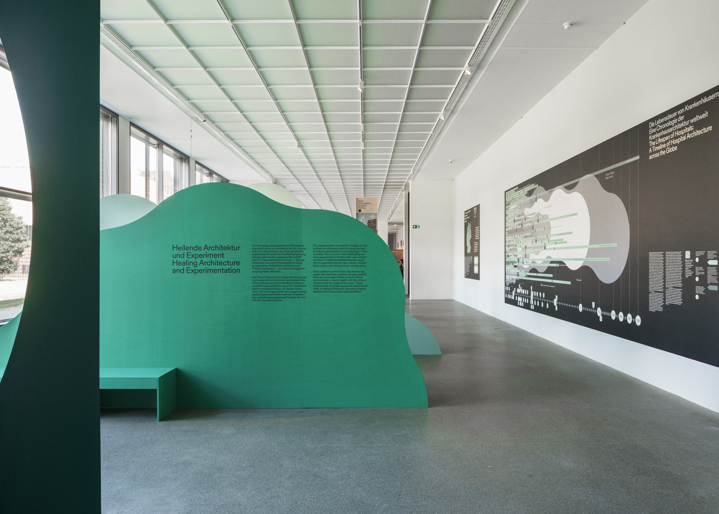 Ein moderner Ausstellungsraum mit grünen, geschwungenen Trennwänden mit Text, einer Bank und großen Fenstern auf der linken Seite, ergänzt durch ein informatives schwarzes Wanddisplay mit dem Titel "automatisch gespeicherter Entwurf", mit weißen Diagrammen und Text auf der rechten Seite. at Hochschule Coburg