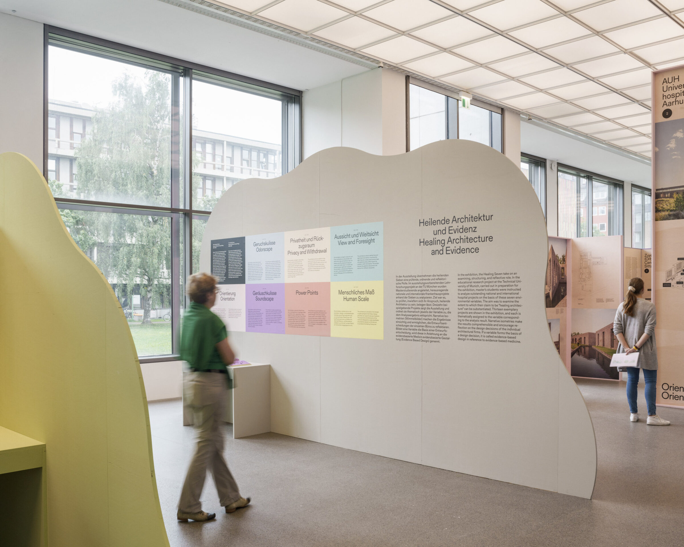 Eine Person betrachtet eine Ausstellung in einem modernen Ausstellungsraum mit großen Fenstern. Bunte Informationstafeln, darunter eine mit der Aufschrift "Automatisch gespeicherter Entwurf", sind an einer gewellten weißen Wand angebracht; ein anderer Besucher geht im Hintergrund. at Hochschule Coburg