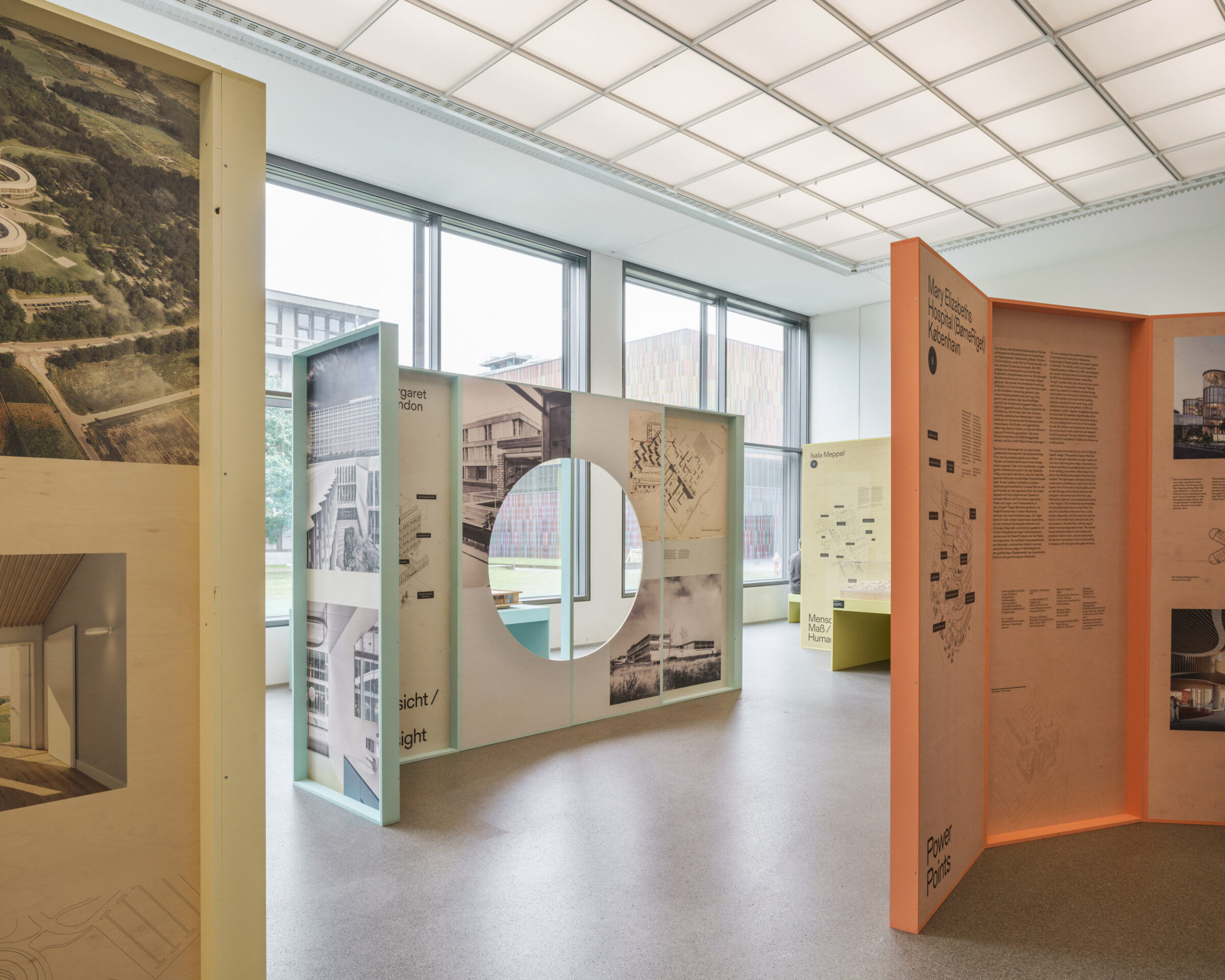 Ein moderner Ausstellungsraum mit Holztafeln, auf denen Architekturzeichnungen, Texte und Fotos zu sehen sind. Eine Tafel hat einen runden Ausschnitt. Große Fenster lassen natürliches Licht herein und geben den Blick frei auf einen grünen Außenbereich - wie ein automatisch gespeicherter Entwurf. at Hochschule Coburg